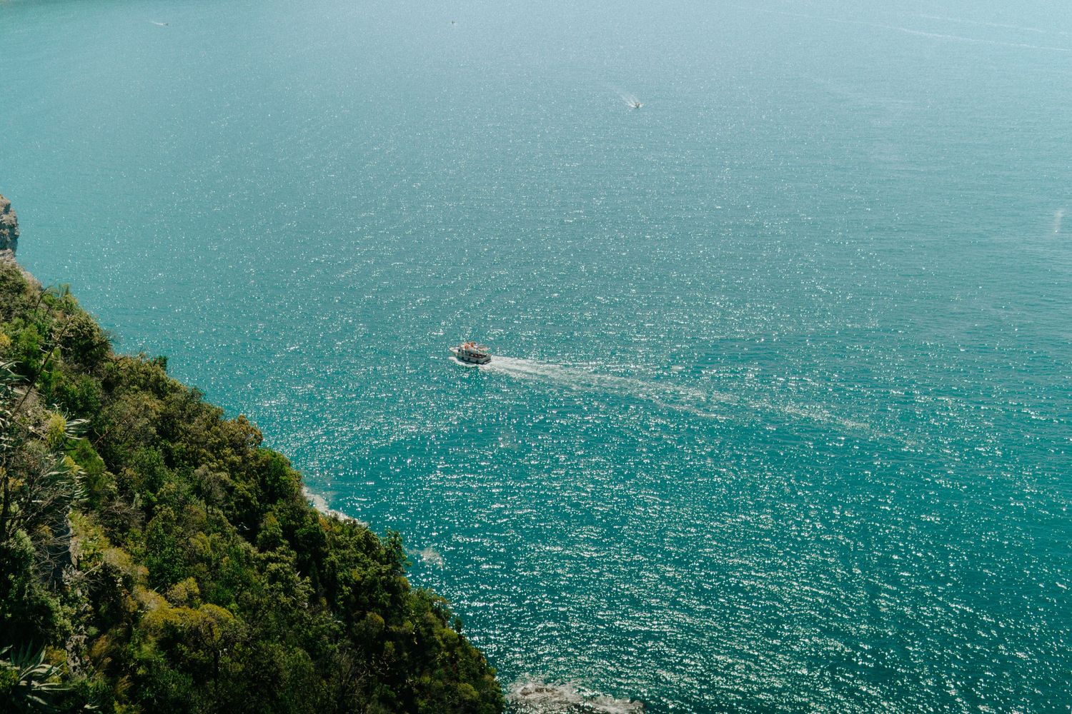 Aerial view of a small boat leaving a wake trail in turquoise Mediterranean waters along a green coastal cliff.