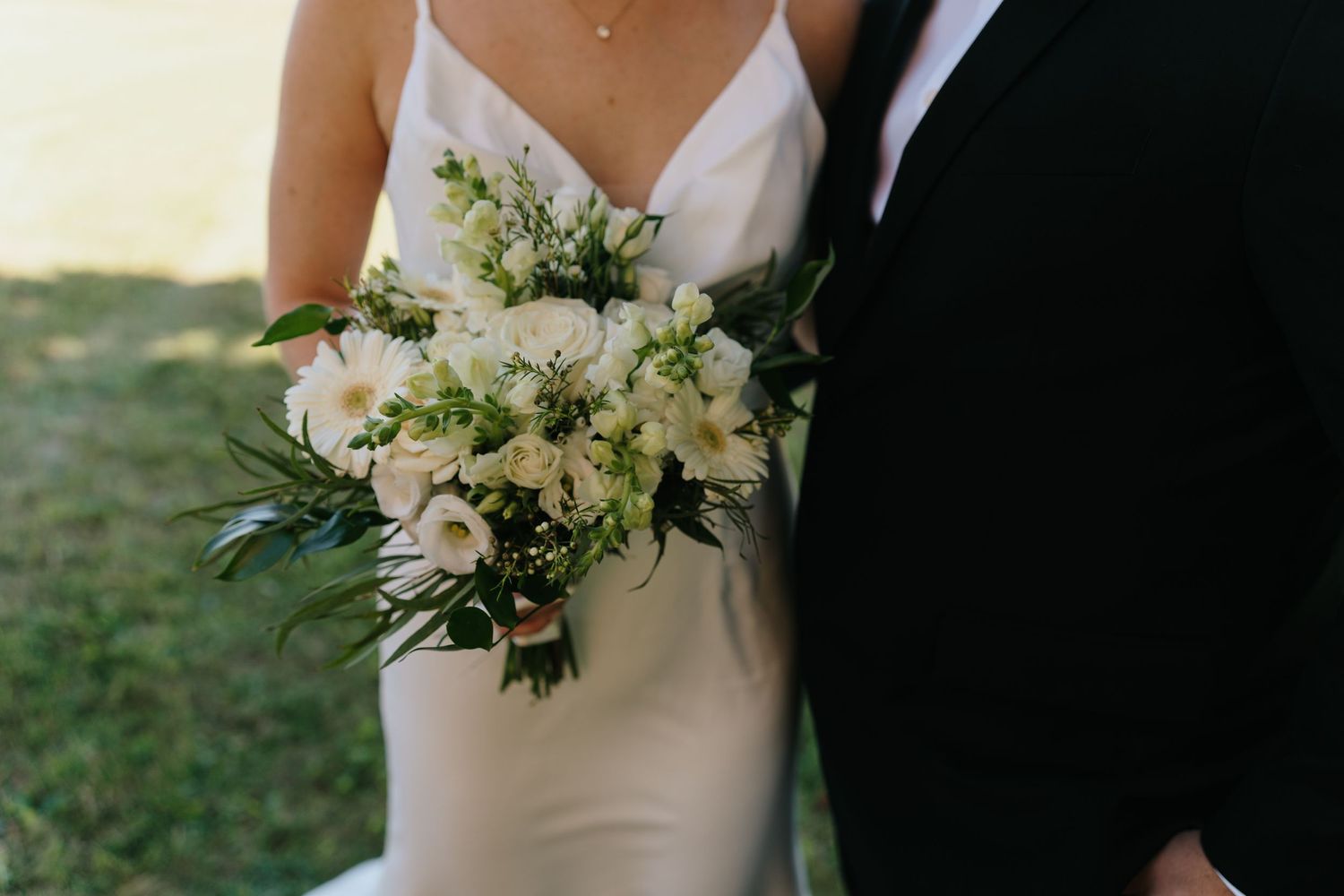 Elegant white and green wedding bouquet held between bride in white dress and groom in black suit.