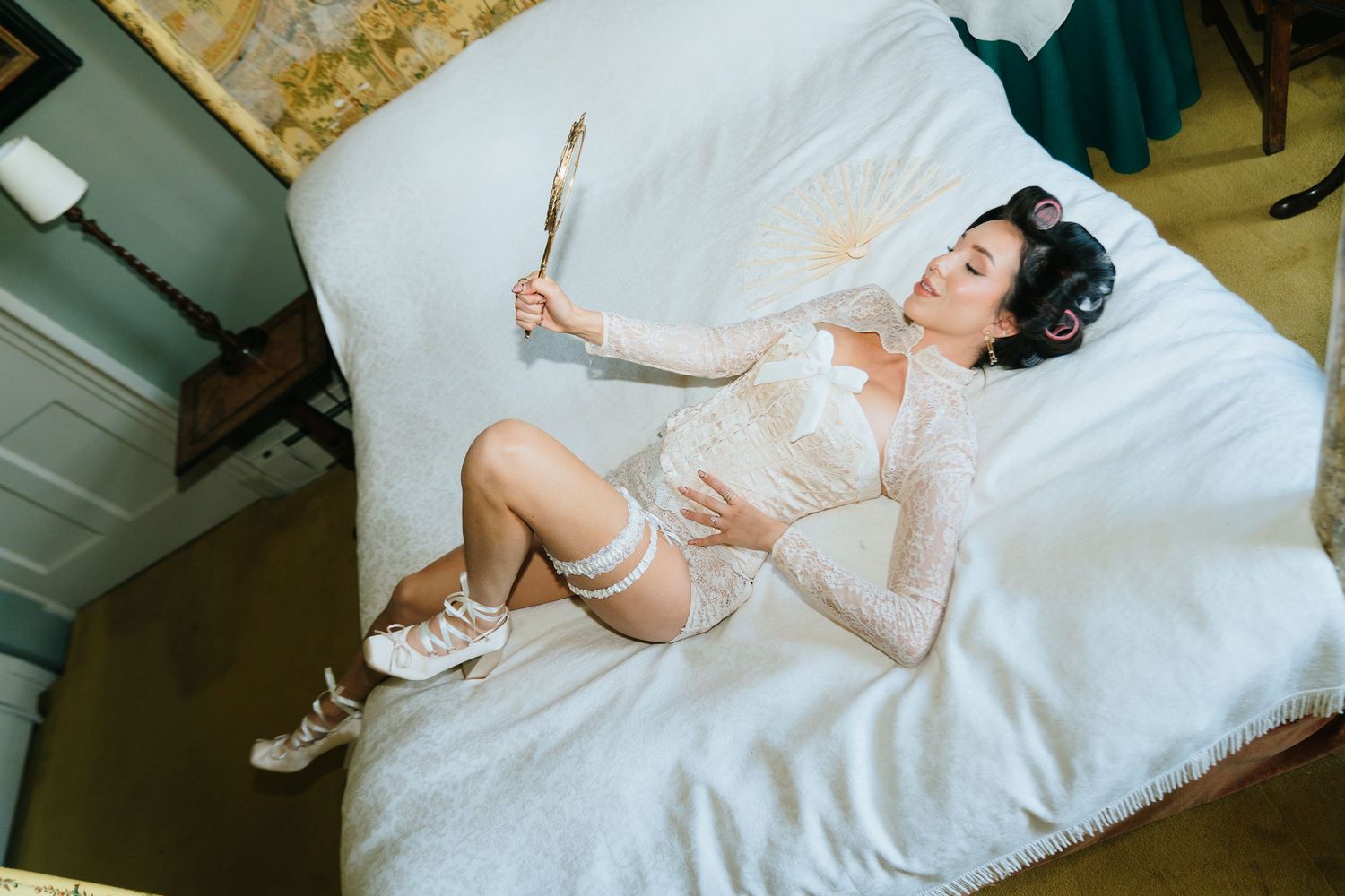 Bride in white lace bodysuit lying on bed holding mirror, wearing garter and heels on wedding day.
