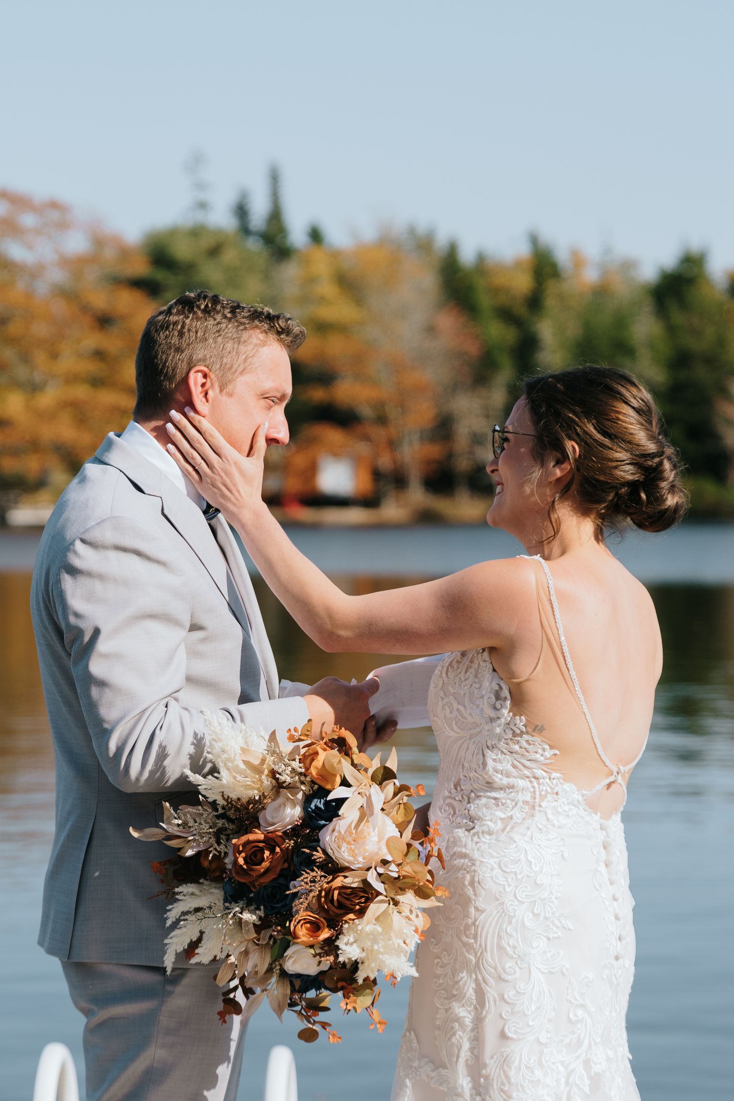 A tender wedding moment by a lake with autumn trees as newlyweds share an intimate touch and hold a fall-themed bouquet.