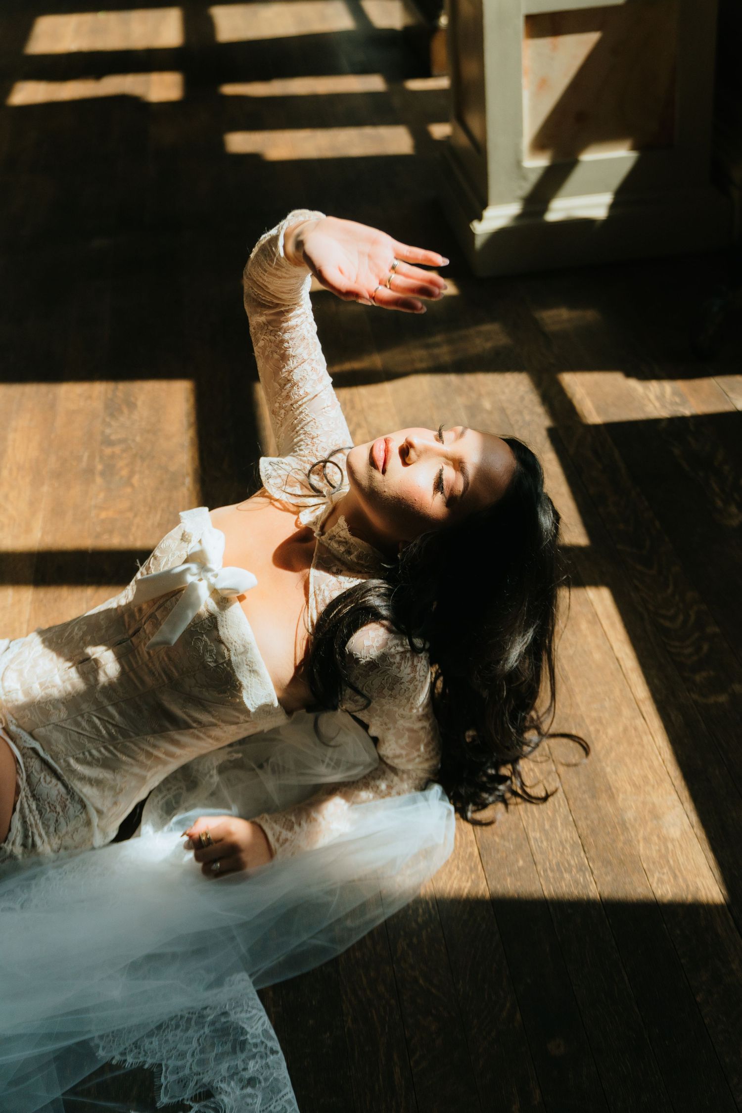 Bride in long-sleeve lace bridal gown posing dramatically on sunlit hardwood floor with arm raised.