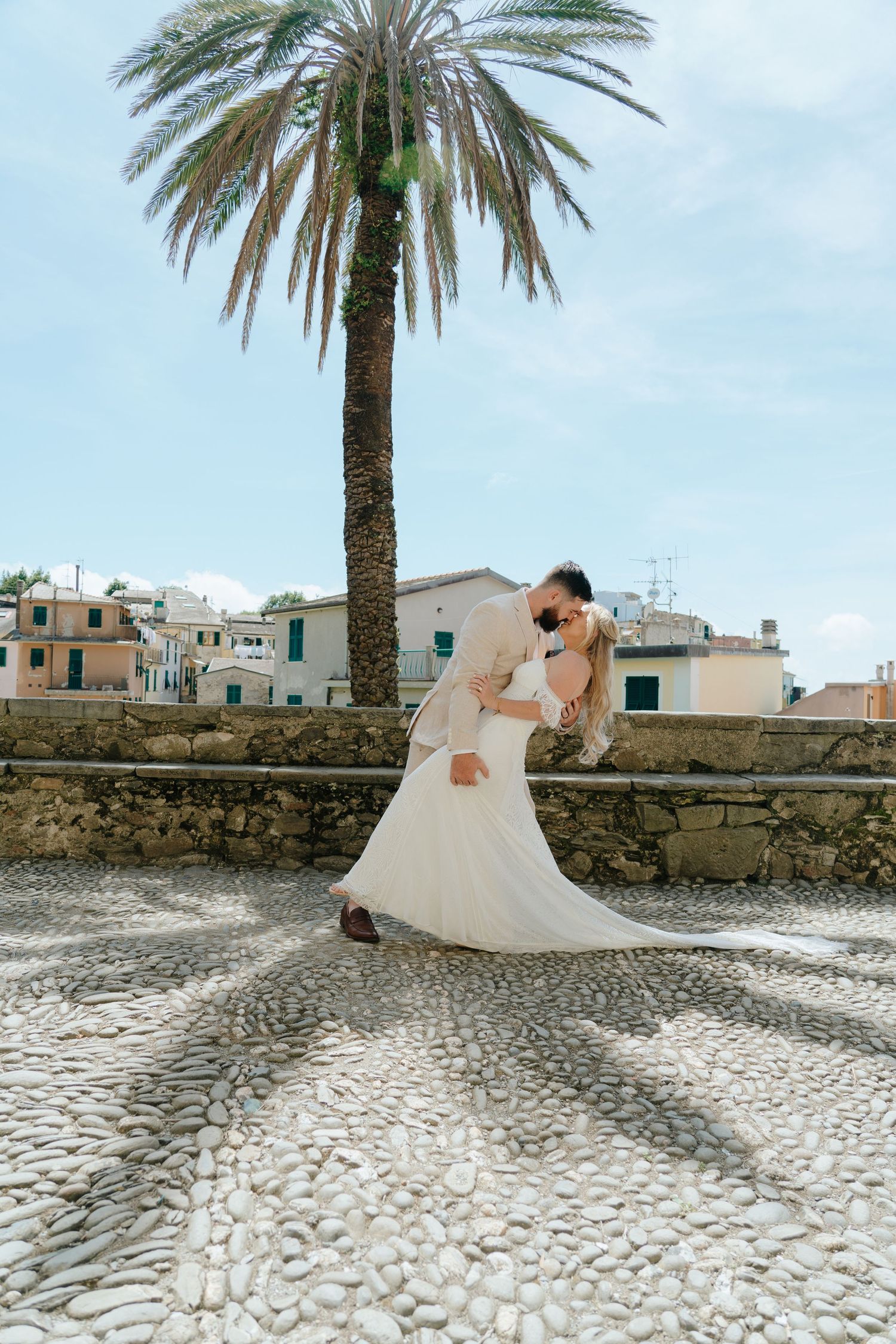 A bride in a flowing white dress poses near a palm tree and stone wall in a Mediterranean setting.