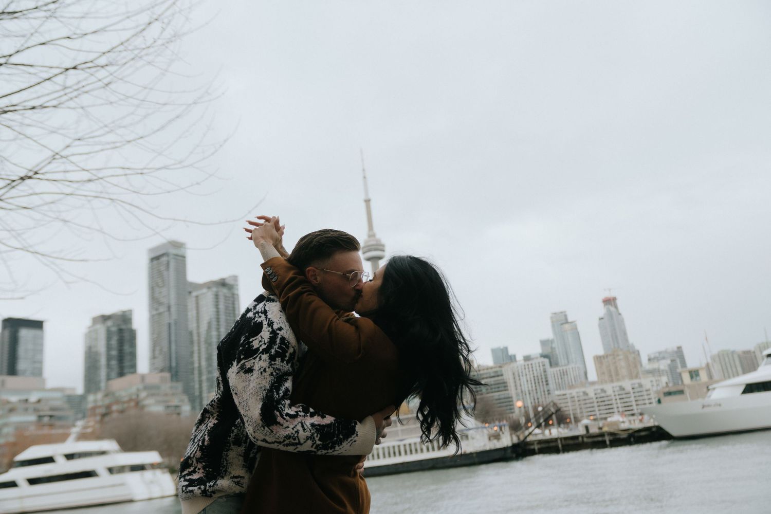 Couple sharing a romantic kiss by the waterfront with Toronto city skyline and CN Tower in the background.