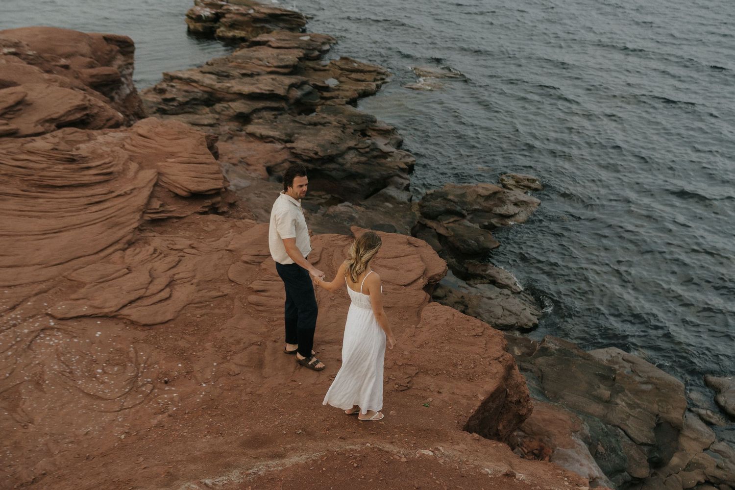A couple stands on red rocky cliffs overlooking dark ocean waters during a coastal photoshoot.