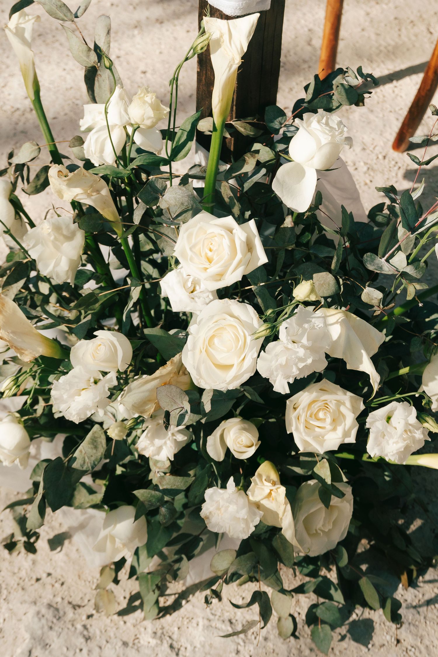 Elegant wedding floral arrangement featuring white roses and greenery on sandy ground.
