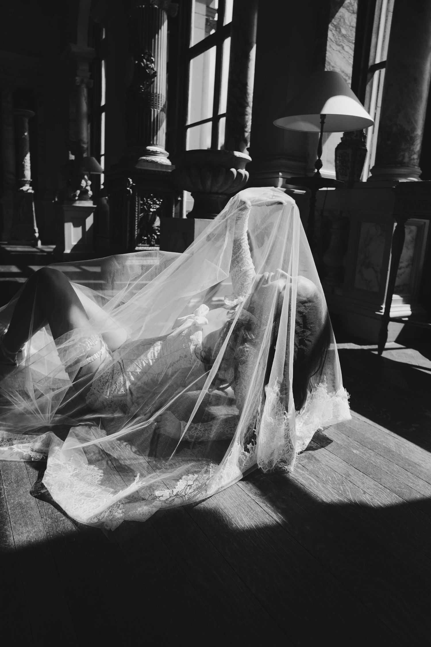 Black and white photo of bride draped in flowing wedding veil on floor bathed in dramatic window light.