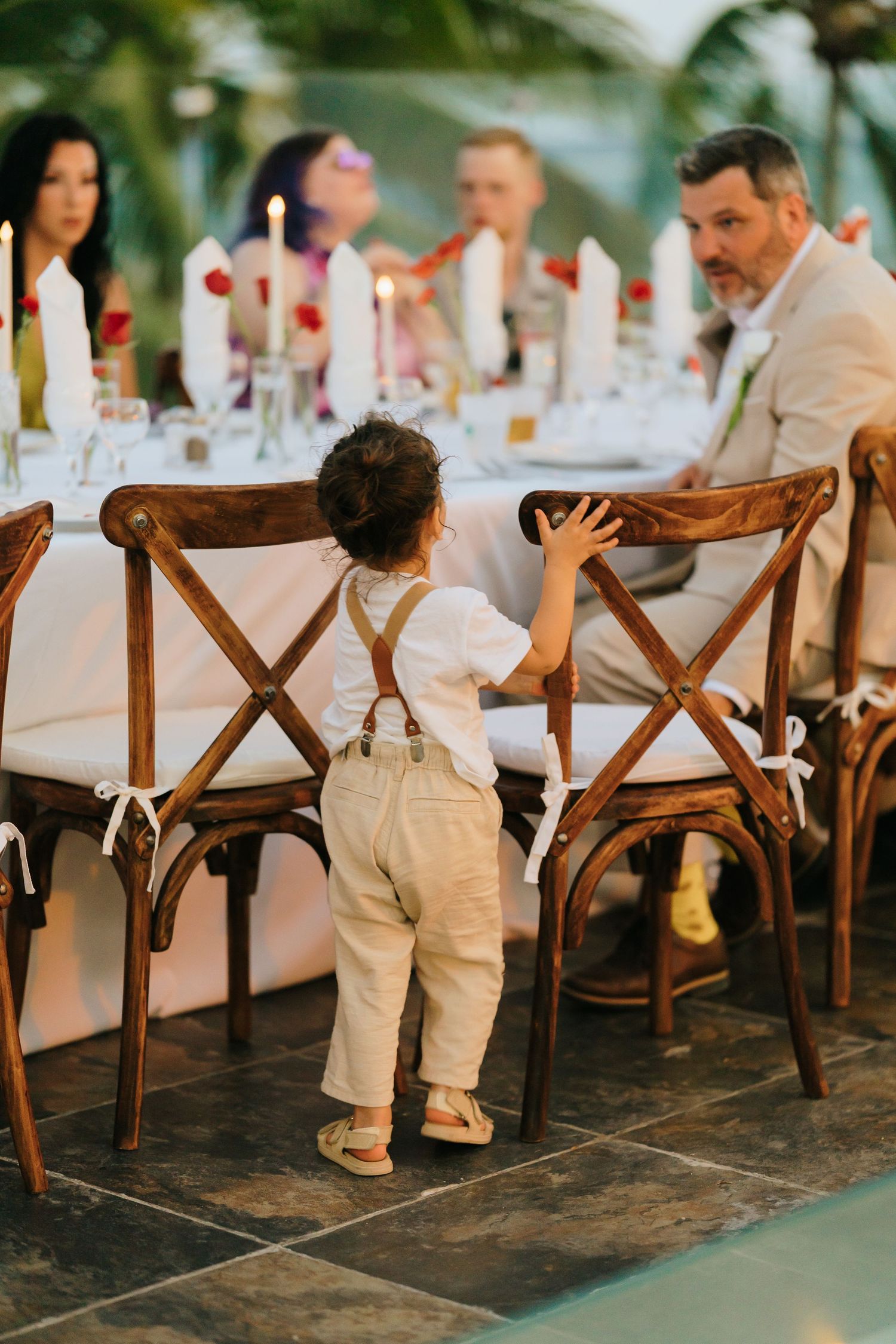 A toddler in suspenders reaches up to a wooden cross-back chair at an elegant outdoor wedding reception dinner table.