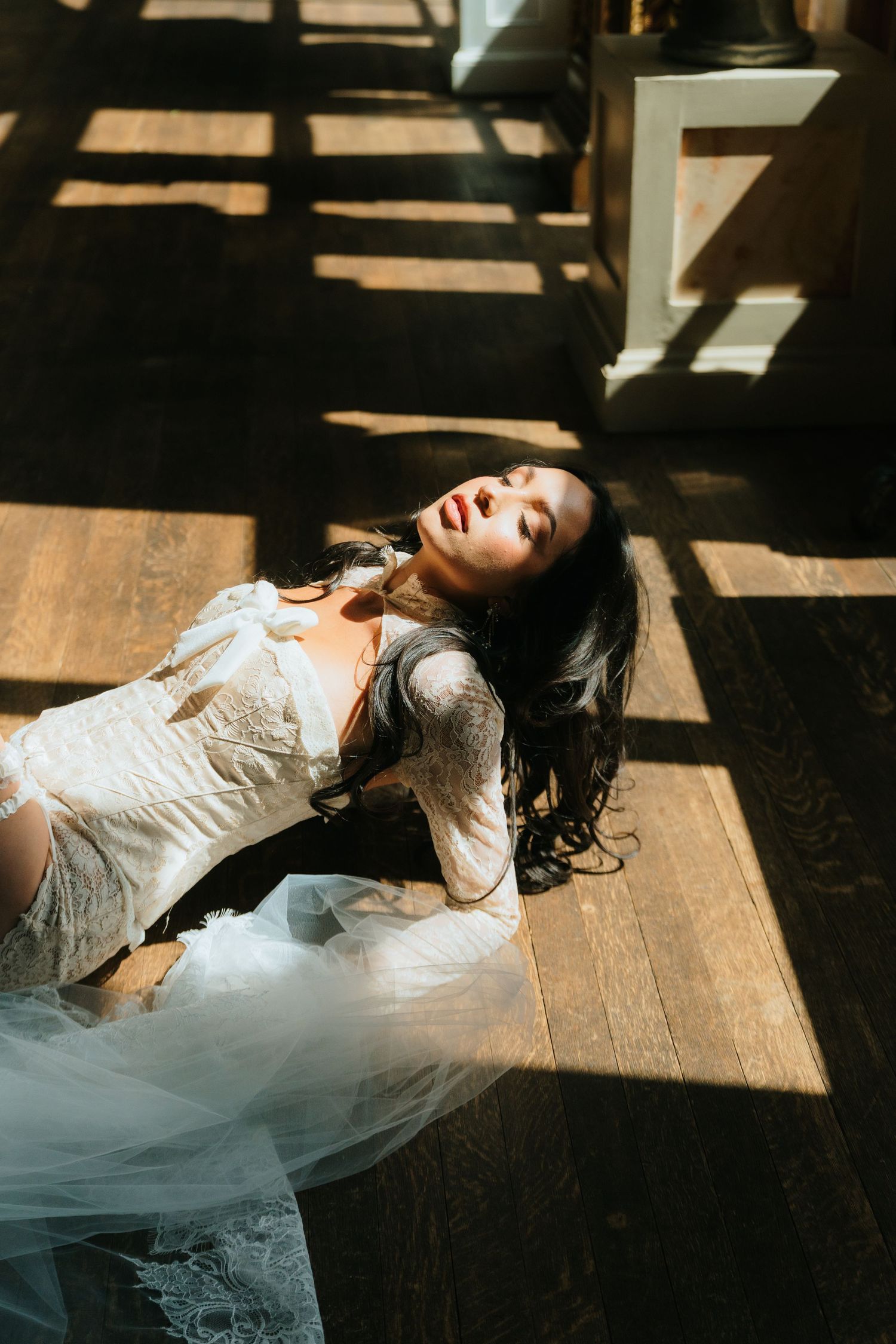 Bride in lace wedding dress lying on hardwood floor bathed in dramatic sunlight and shadow patterns.