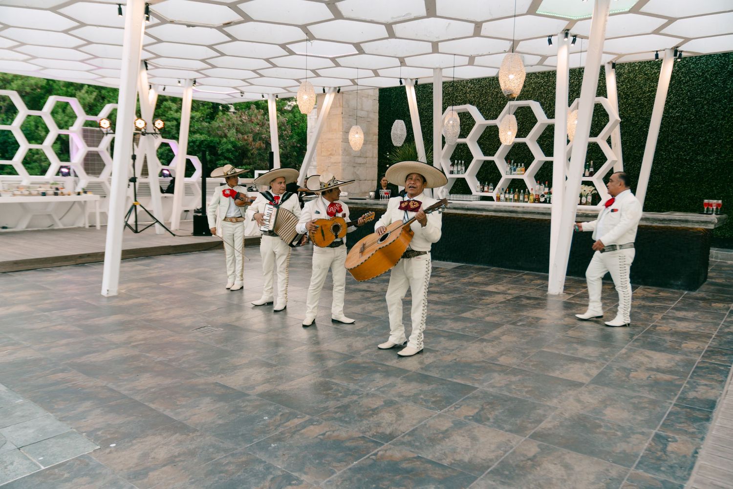 Mariachi band in white suits performing outdoors under a decorative white pergola with honeycomb wall backdrop in Mexico