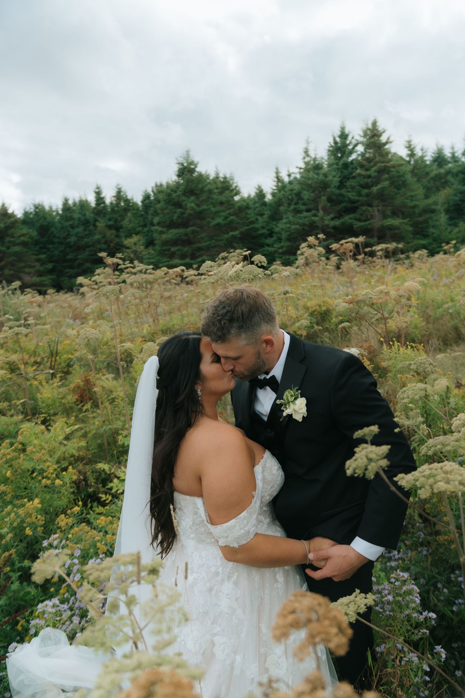 A romantic wedding photo in a wildflower field with evergreen trees in the background.