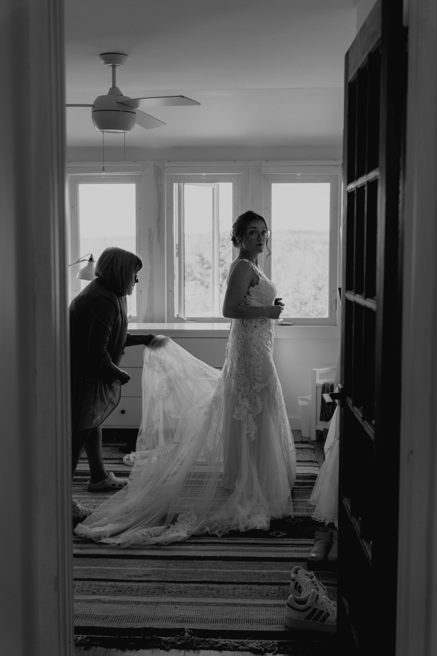 A bride in a lace gown gets ready in a sunlit room while her dress train is arranged.
