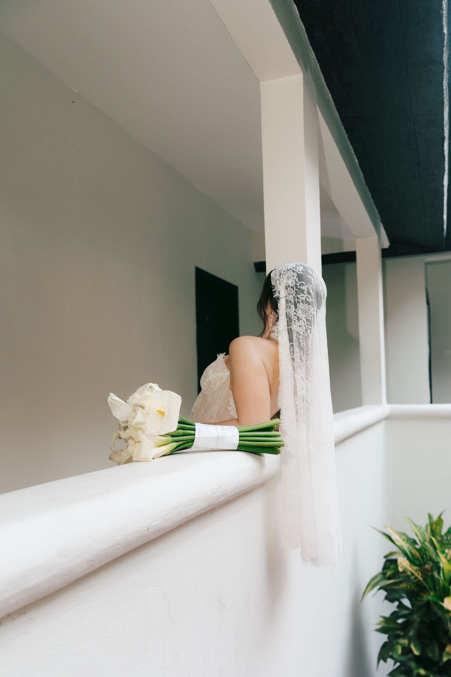 Bride in white dress and veil stands on balcony beside elegant calla lily bouquet on white railing.