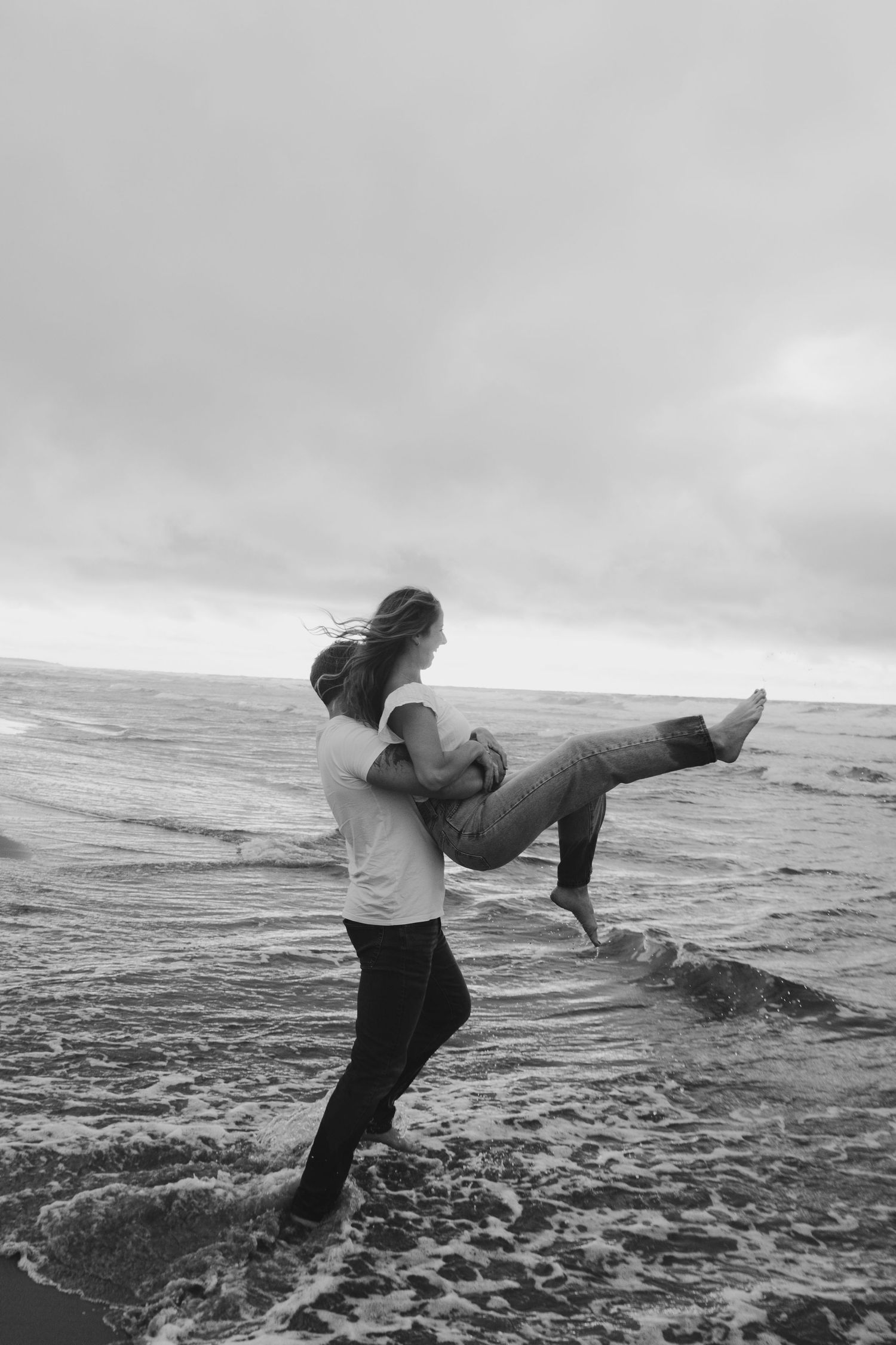 Black and white photo of a couple embracing playfully on a stormy beach, waves crashing around their feet.