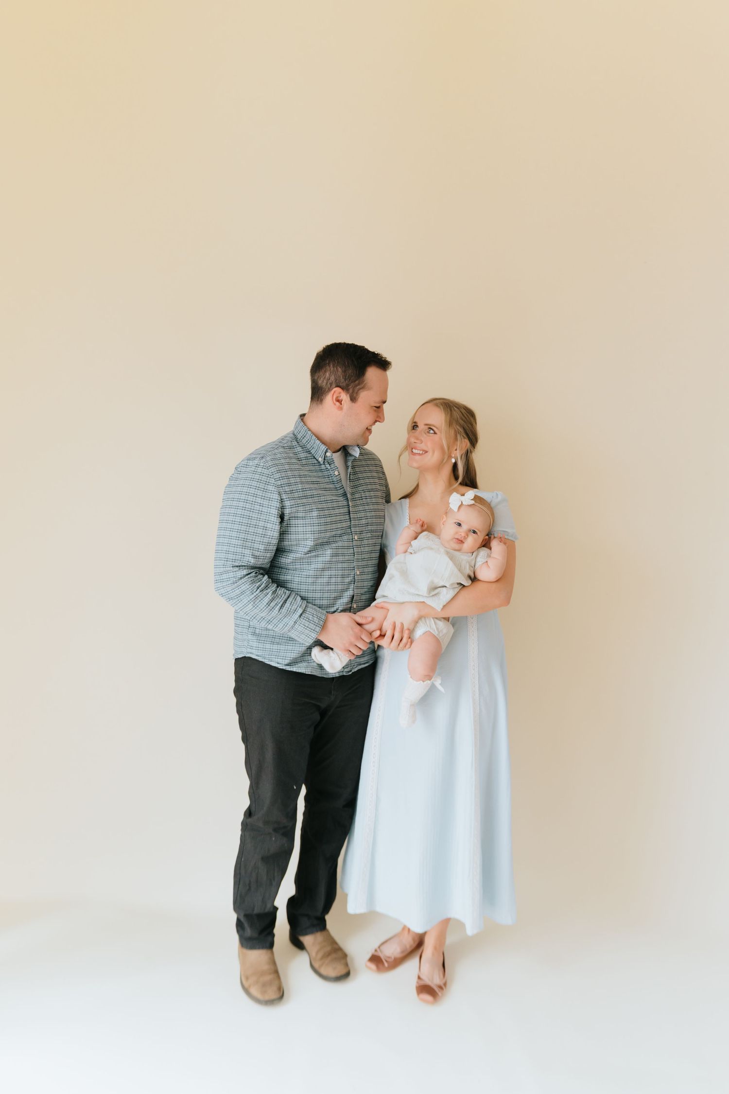 A couple in a bright studio smiles at each other, the woman holding a baby in a white outfit.