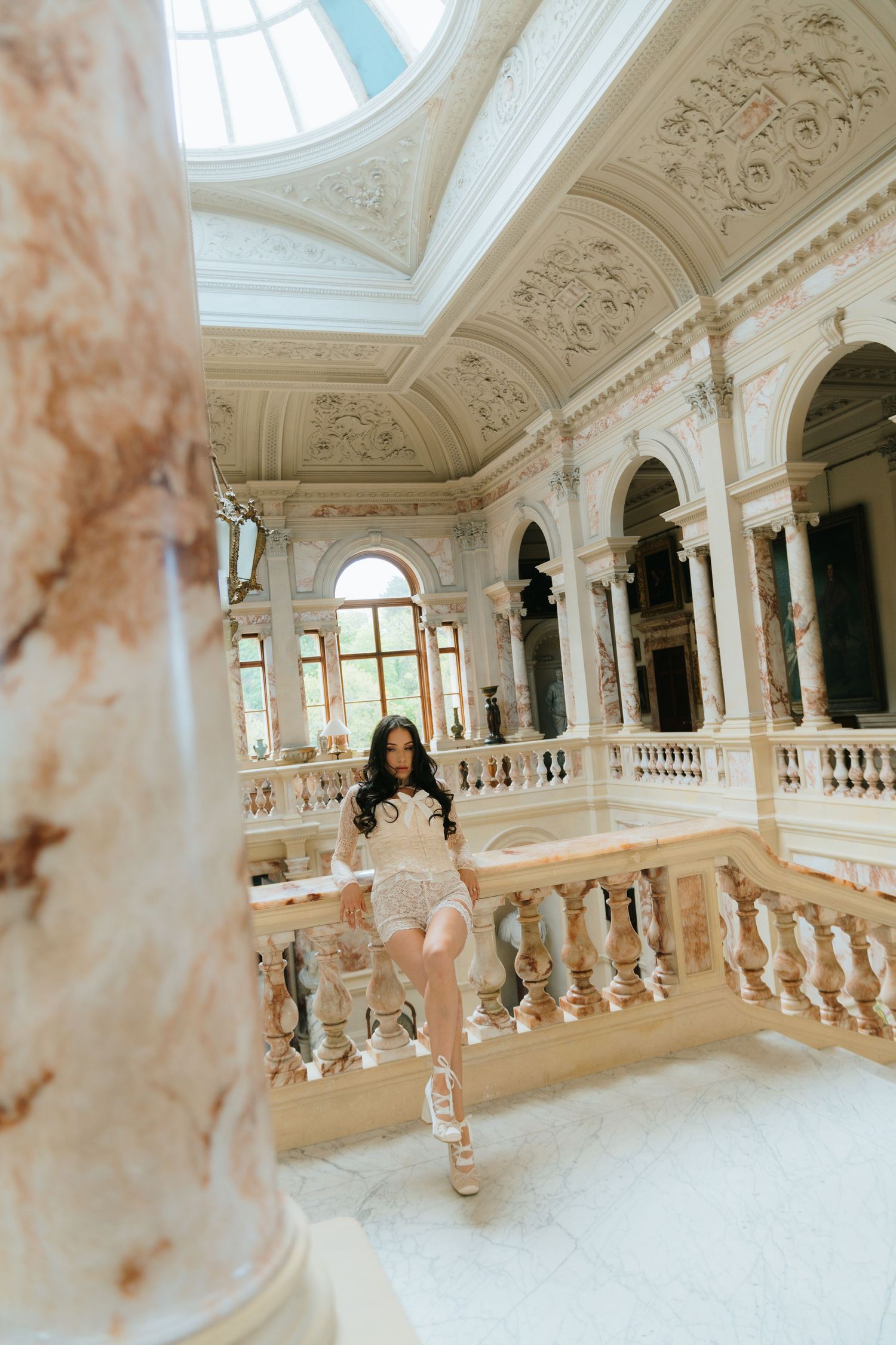 Woman in a short lace outfit posing on marble stairs inside an ornate luxury palace with elaborate ceiling decor.