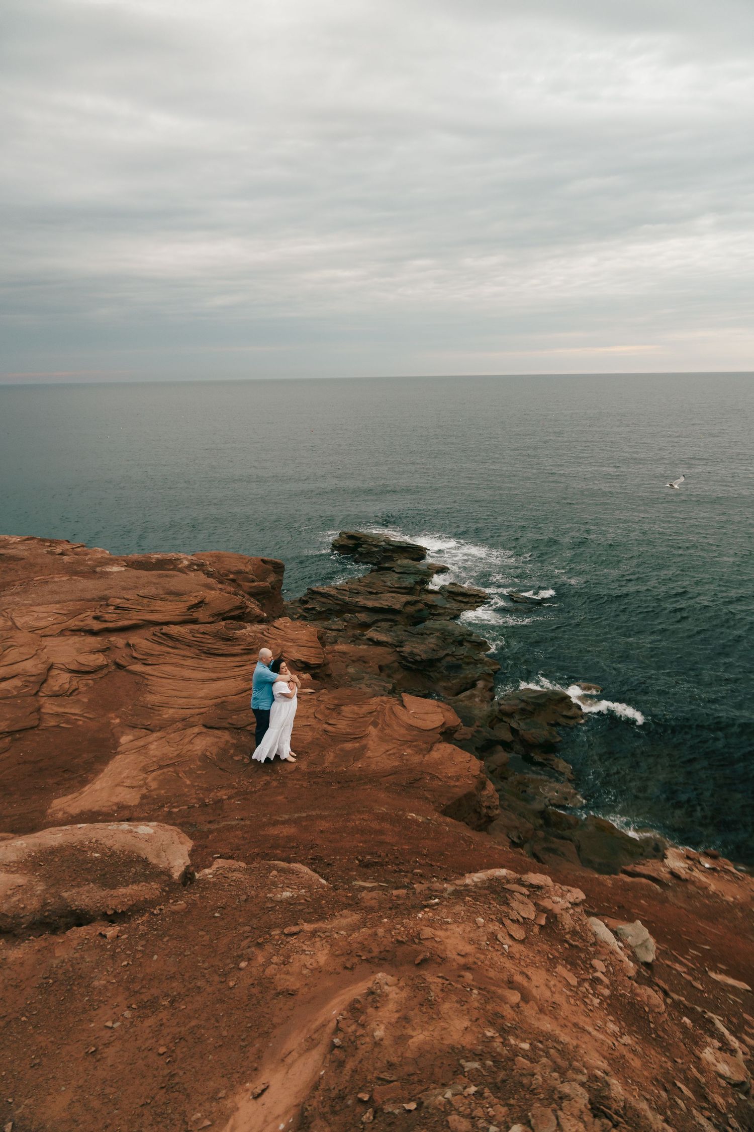 Couple embracing on red rocky coastal cliffs overlooking turquoise ocean waves under cloudy skies.