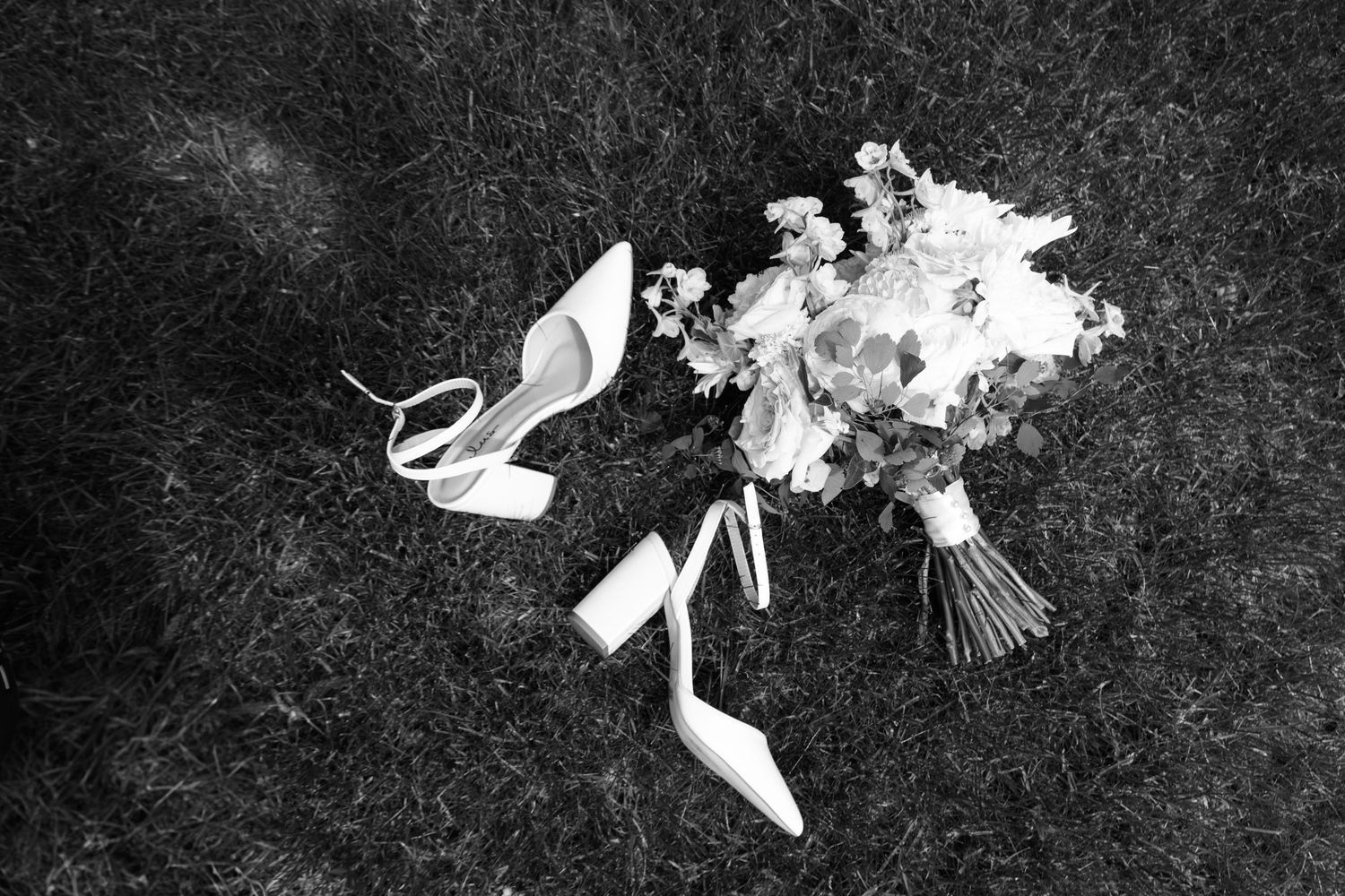 Black and white photo of bridal heels and floral bouquet arranged on grass.
