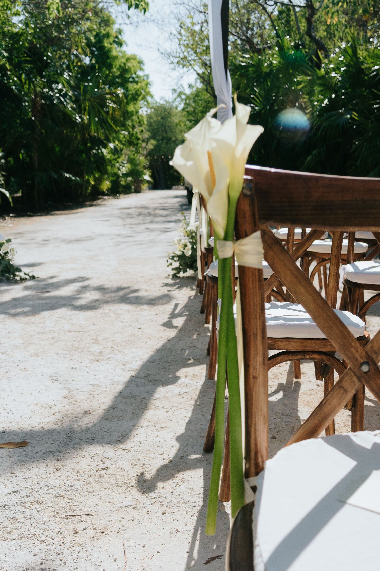 White calla lily floral arrangement on wooden wedding ceremony chairs lining a sandy outdoor aisle surrounded by lush greenery.