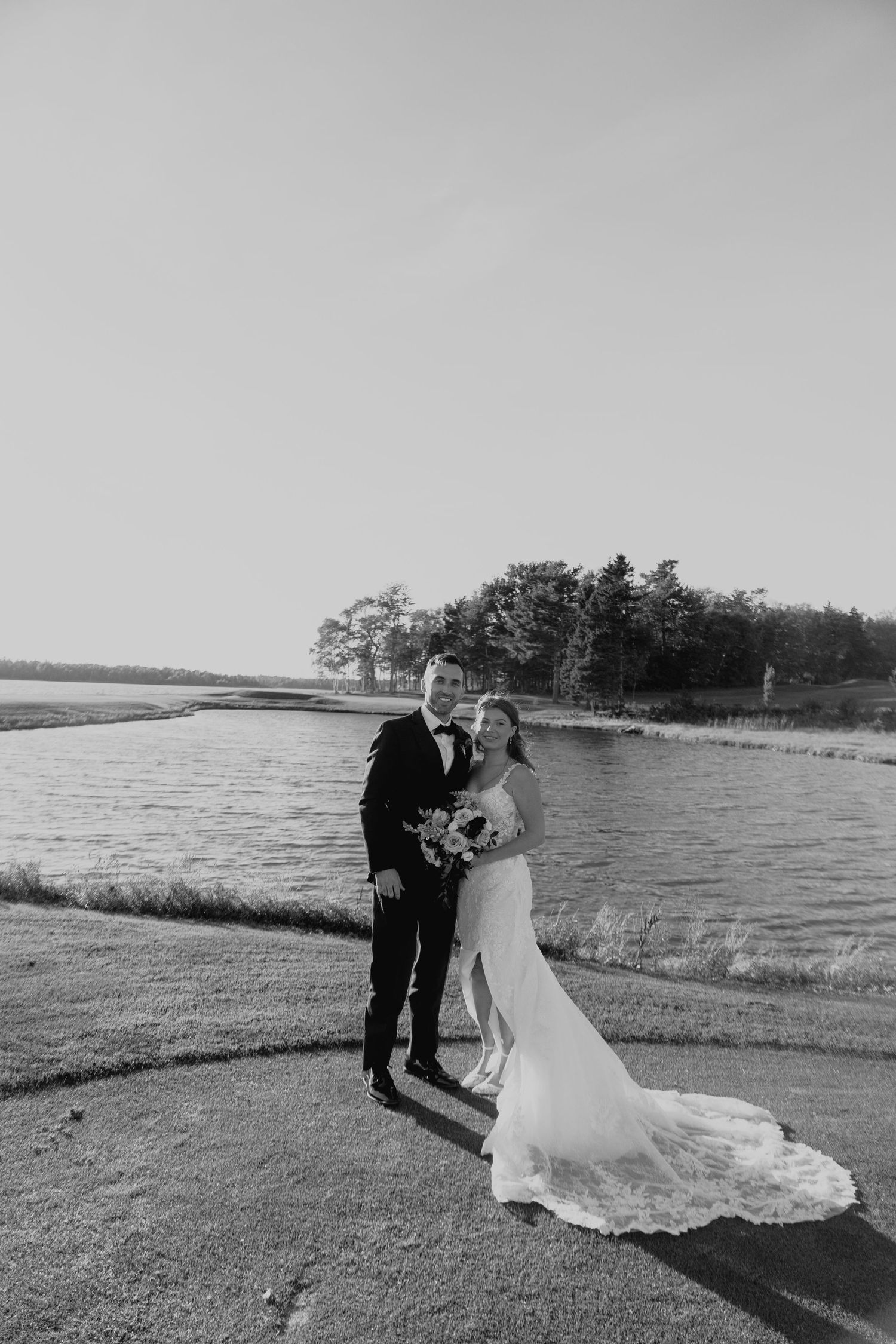 Black and white photo of a bride and groom posing by a serene waterfront, bride in lace gown holding bouquet.