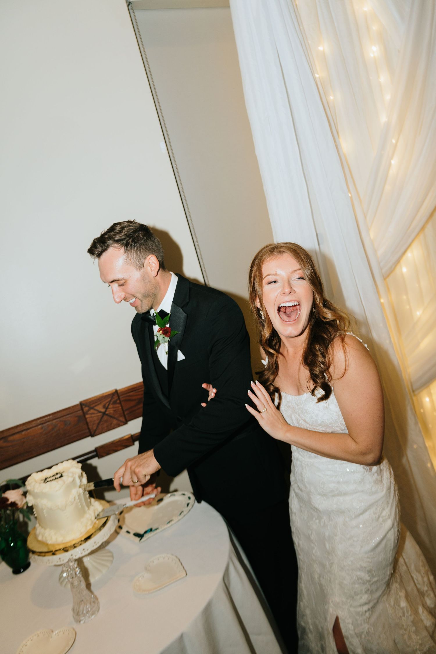 Bride laughing joyfully while groom cuts wedding cake at reception, bride in lace gown, groom in black tuxedo.
