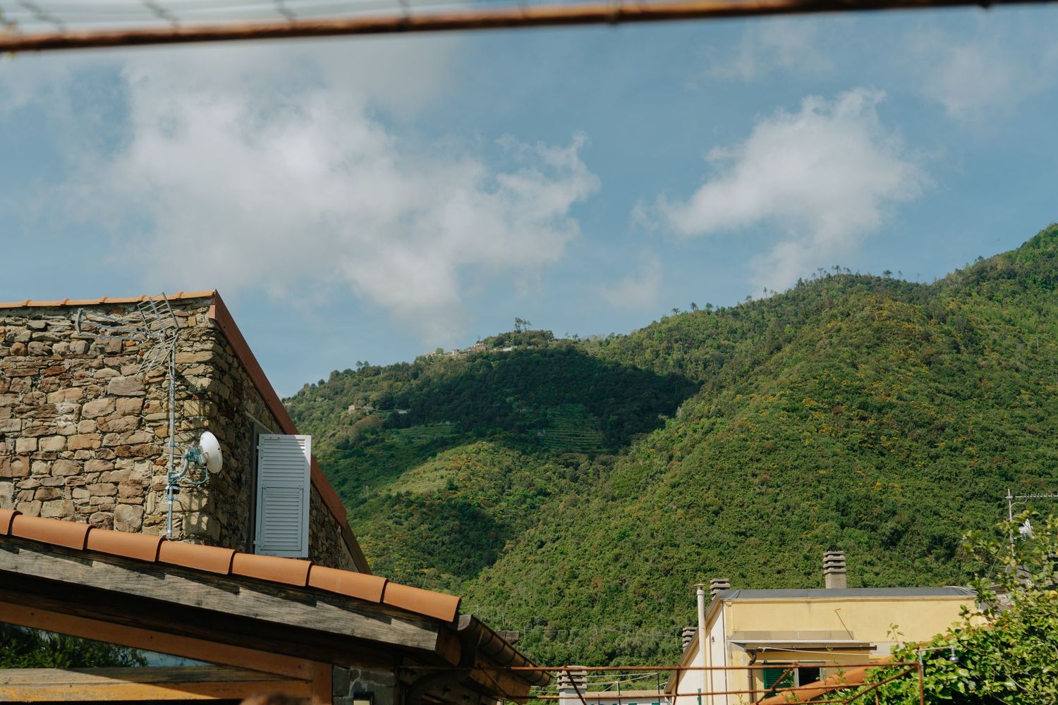 Stone building with terracotta roof tiles against green mountain landscape under blue sky with white clouds.