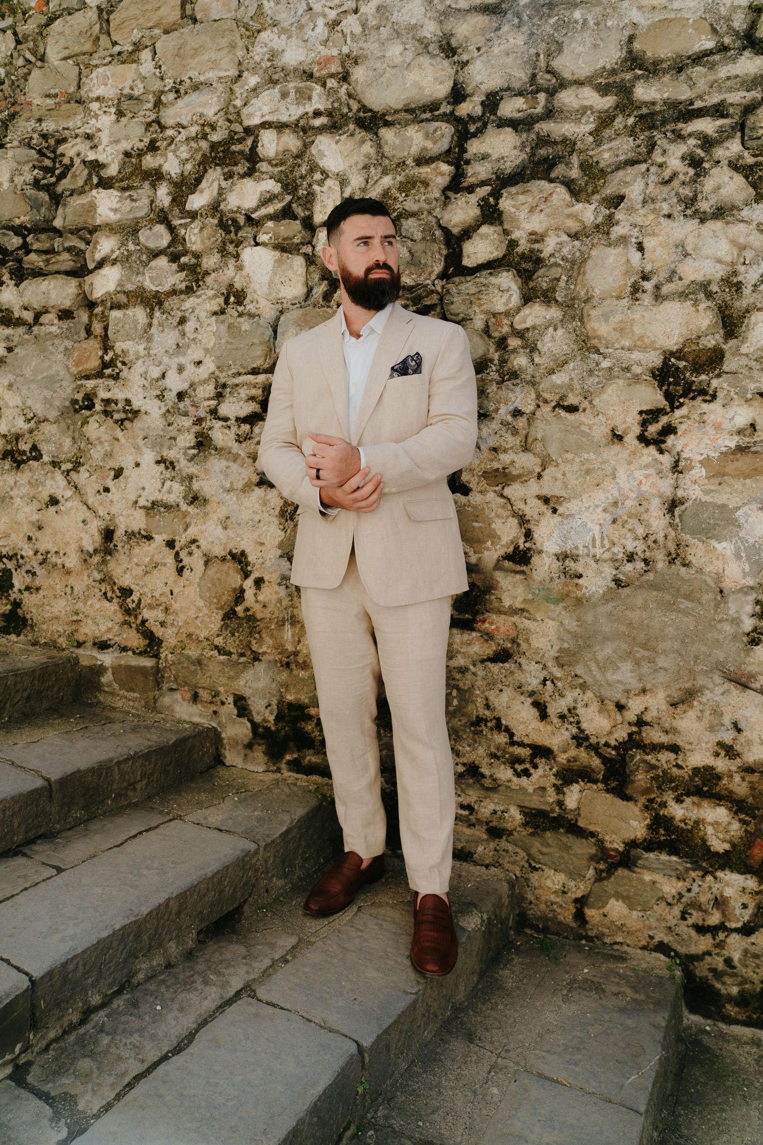 A groom in a beige suit and brown shoes stands on stone steps against an old stone wall.