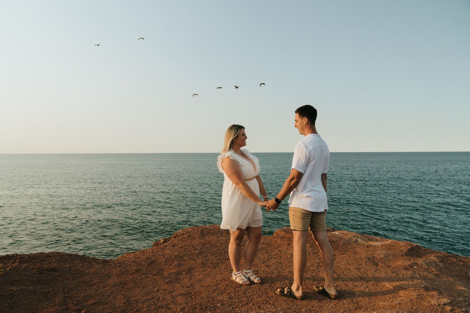 A couple in white outfits stands at the edge of a rocky cliff overlooking a serene ocean view at sunset.