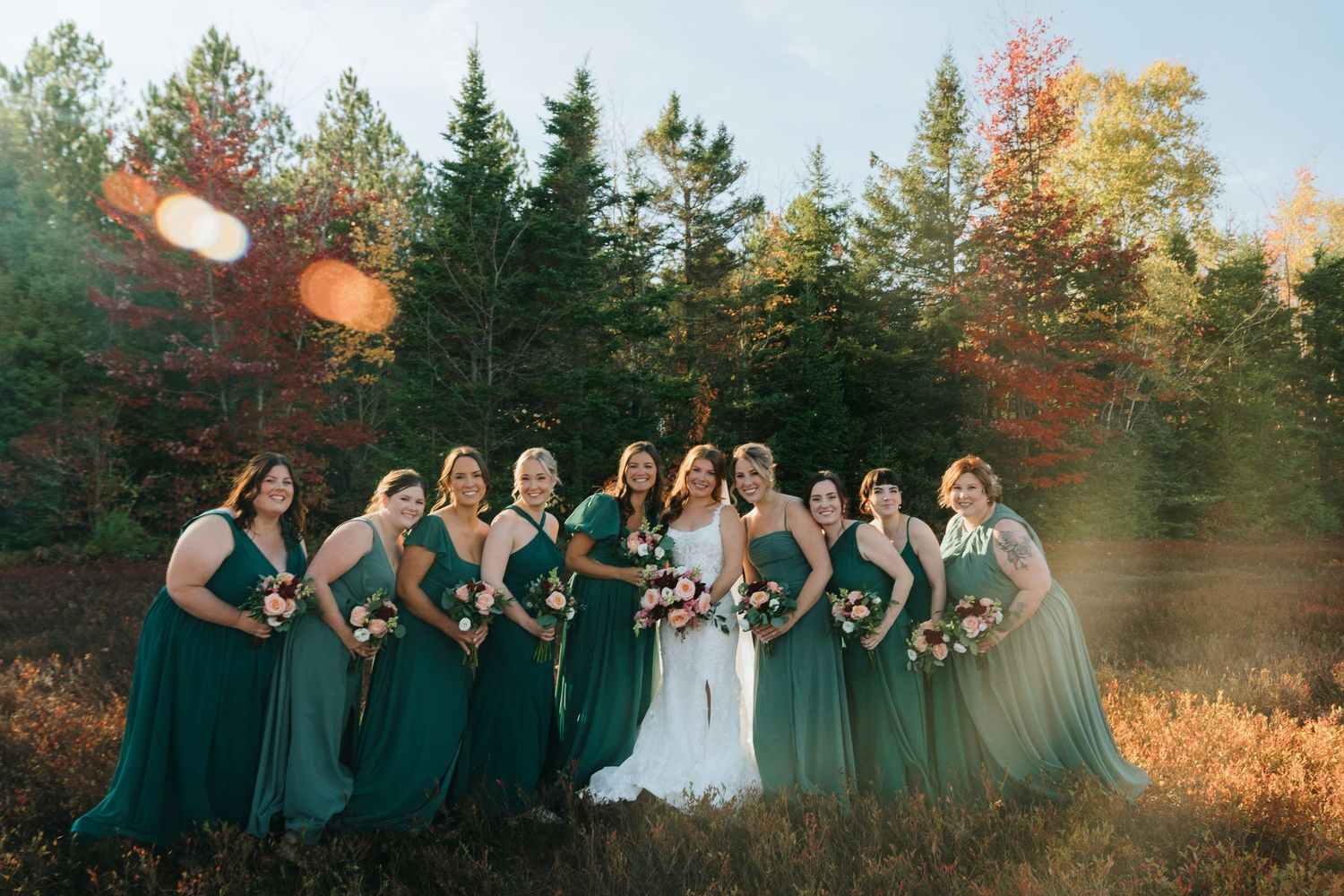 Bride poses with bridesmaids in emerald green dresses surrounded by autumn foliage and pine trees.