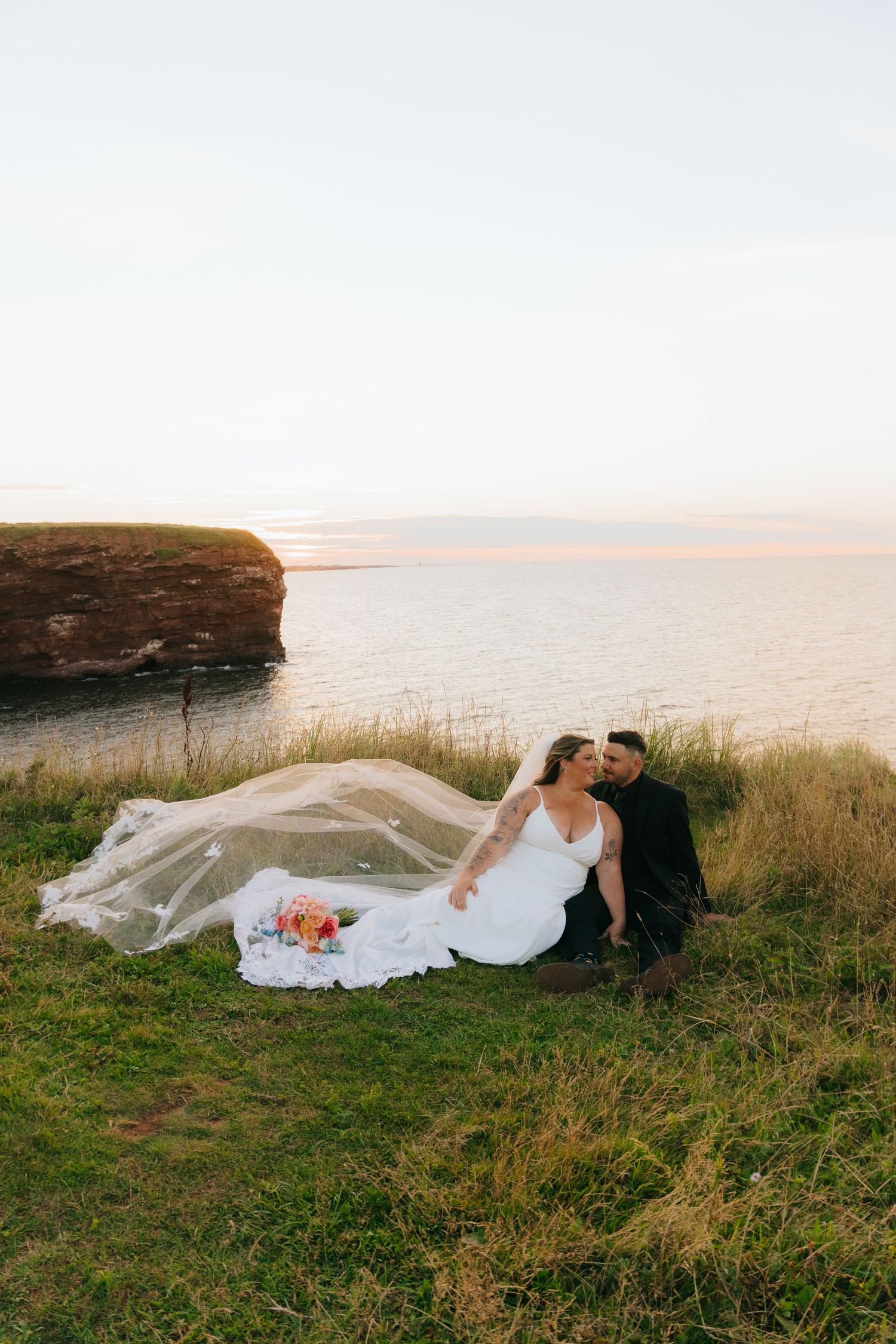 Bride and groom sit on coastal cliffside at sunset, veil flowing in the wind, with red rock formations and calm sea behind.