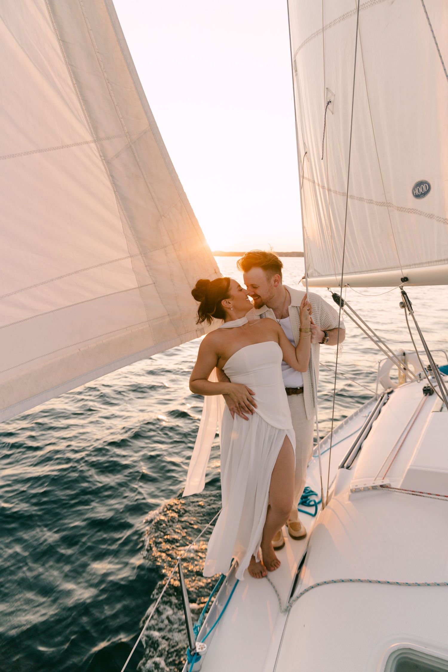 Romantic couple embracing on a sailboat deck at golden sunset, woman in white dress, man in casual attire.