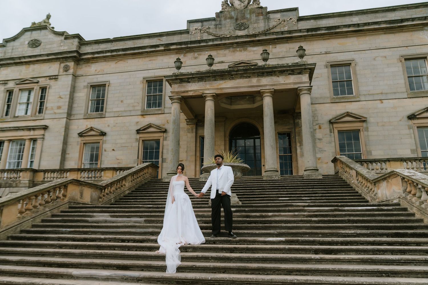 A bride and groom hold hands on grand stone steps of a historic mansion venue on their wedding day.
