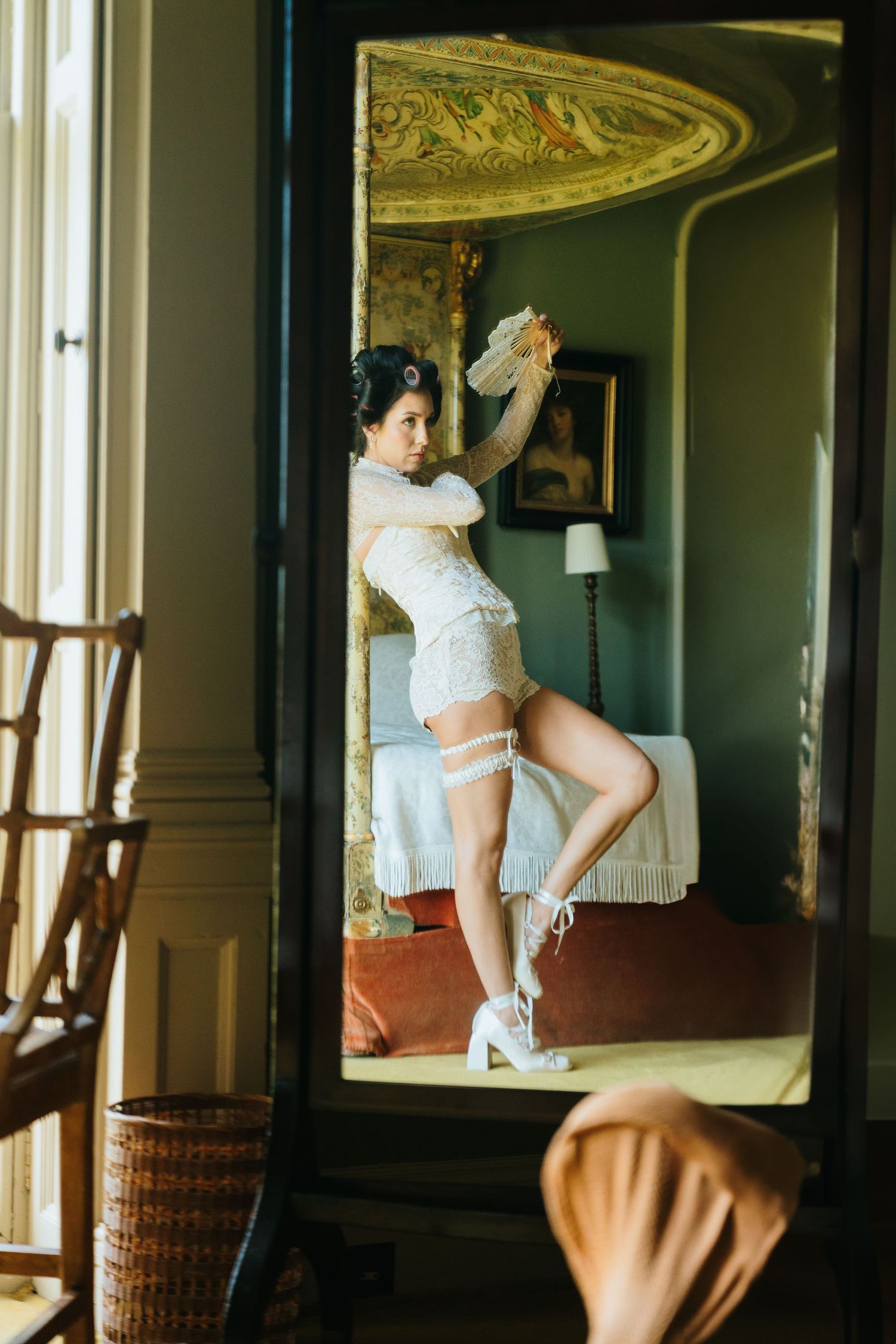 Bride in white lace lingerie and heels poses elegantly before an ornate mirror in a vintage bedroom.