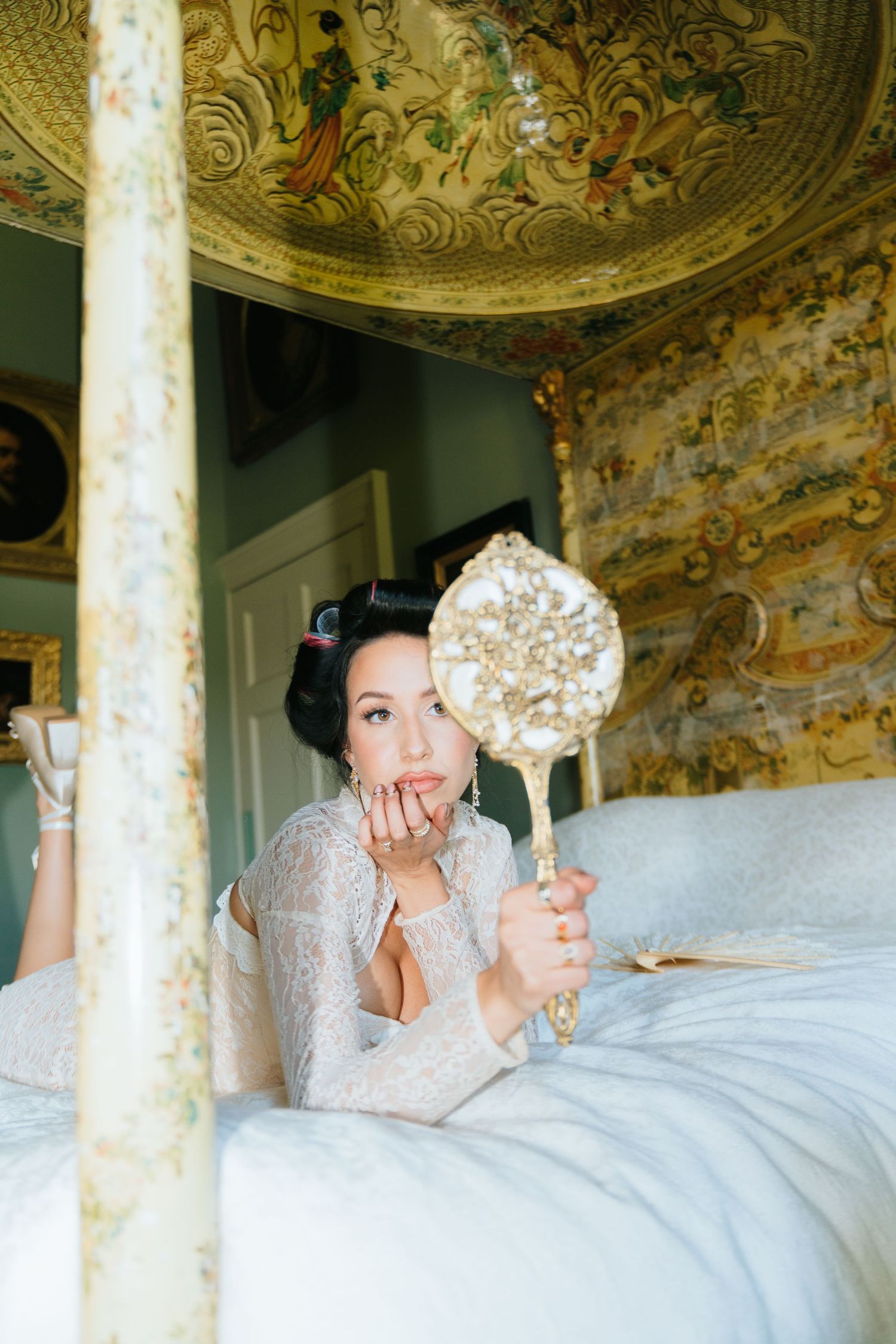 Dark-haired bride in white lace robe lying on bed holding vintage gold hand mirror.