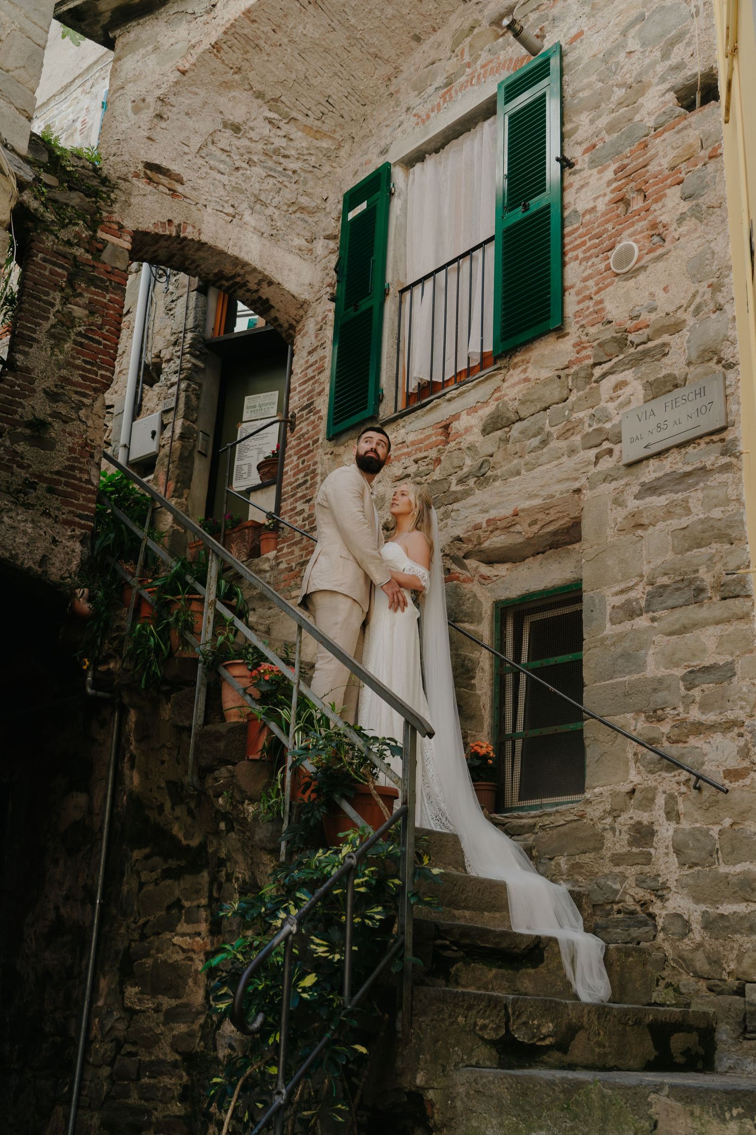 A couple stands on rustic stone steps of an old Italian building with green shutters and weathered walls.