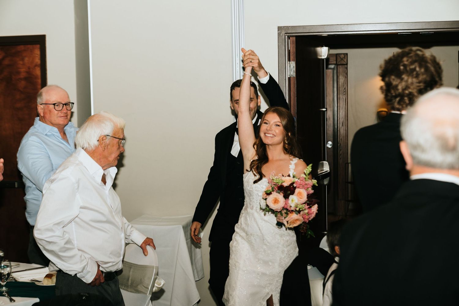 Bride and groom make grand entrance holding hands raised at elegant wedding reception with seated guests.