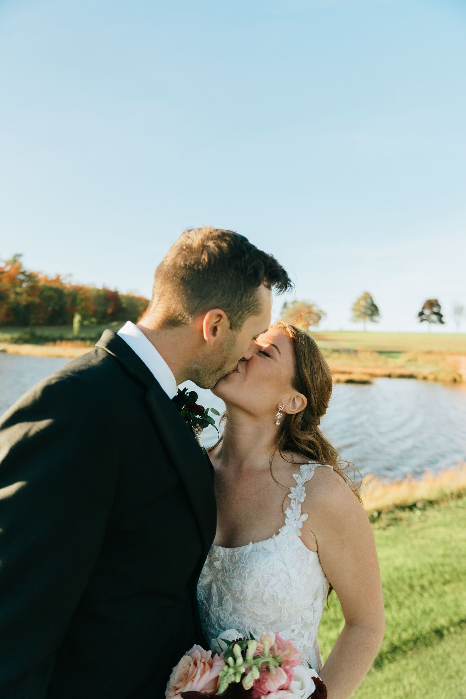 Bride and groom share a romantic kiss outdoors near a pond on their wedding day, with fall foliage in the background.