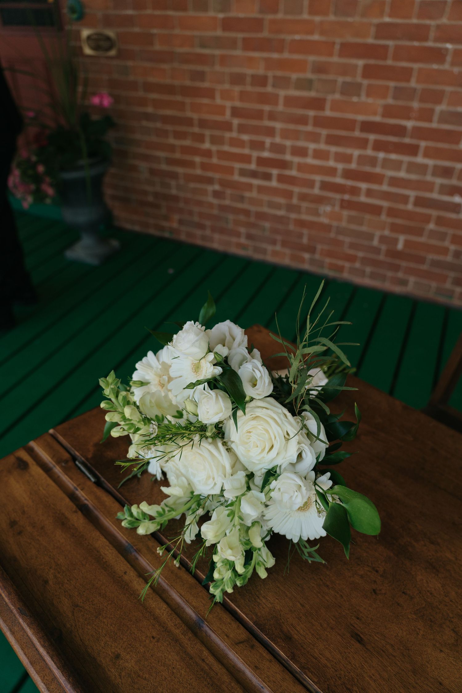 White floral bouquet with roses and greenery resting on a rustic wooden surface against a brick wall background.