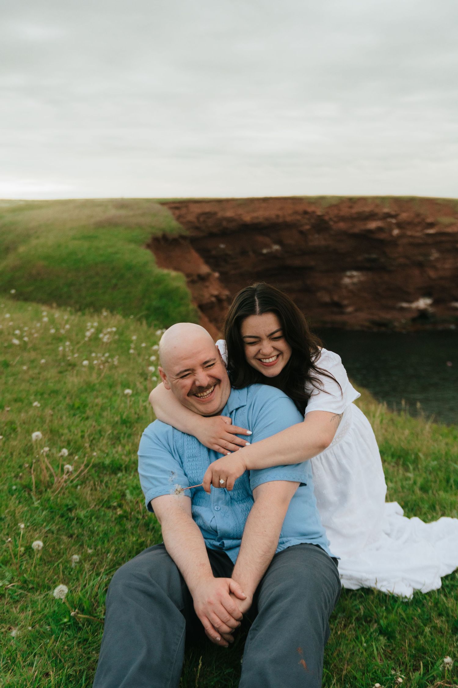 A laughing couple sits on green cliffside grass, woman in white dress embracing bald man in blue shirt.