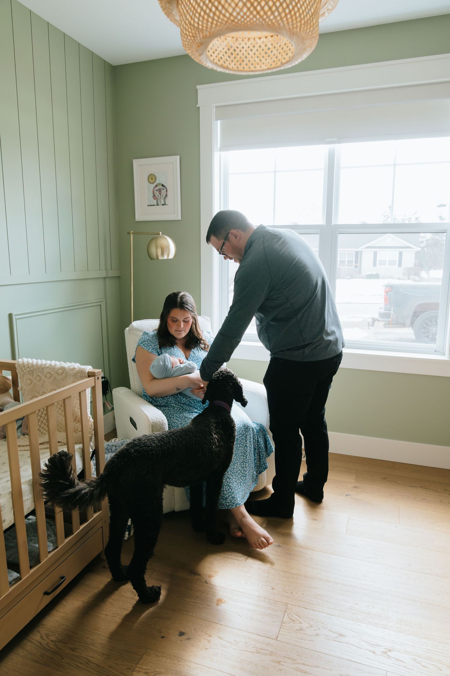 A family with a newborn sits in a sage green nursery with a black dog, wooden crib, and warm lighting.