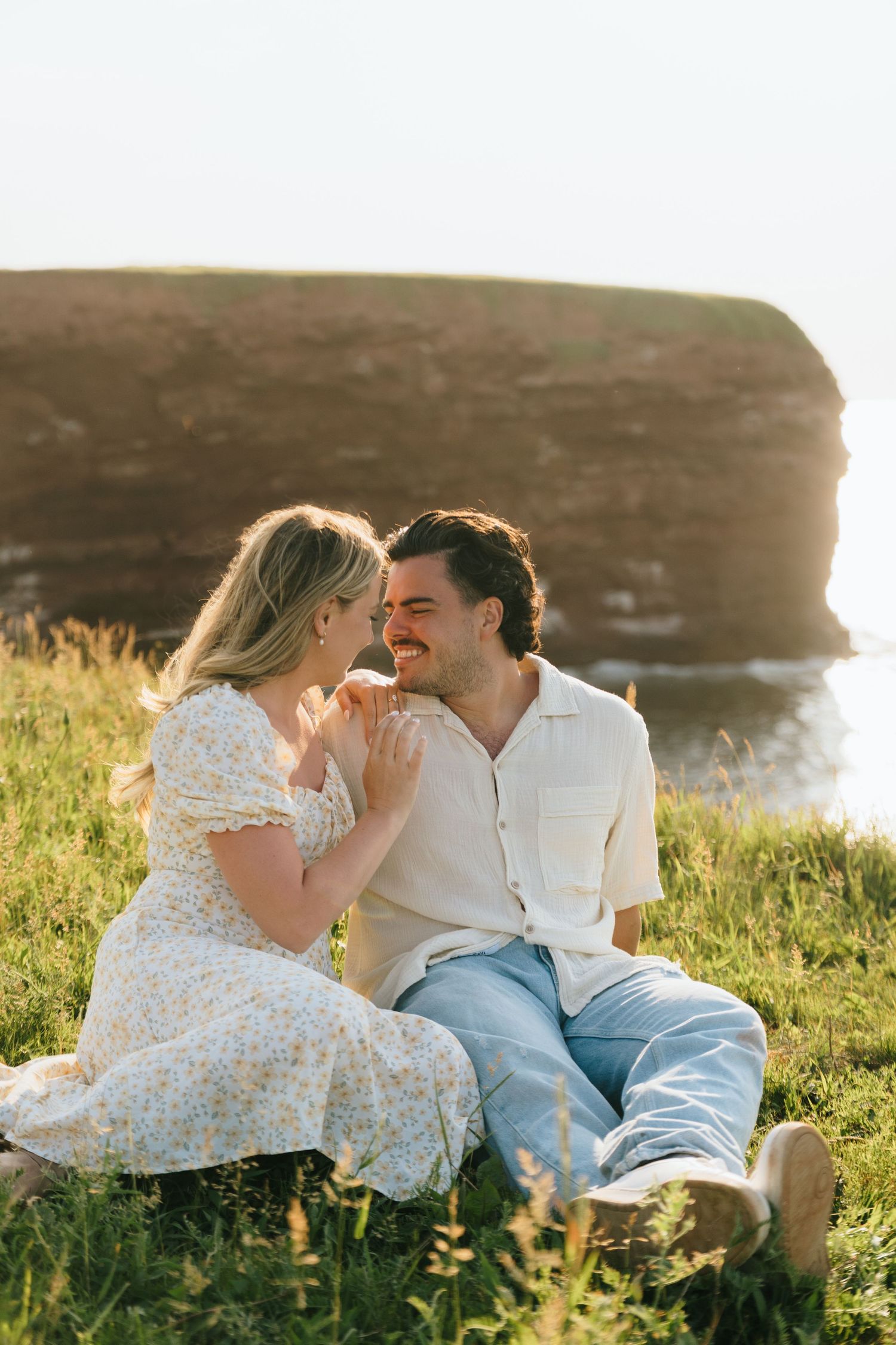 Couple sharing a romantic moment while sitting in a grassy field at sunset with cliffs in the background in Cape Tryon, PEI