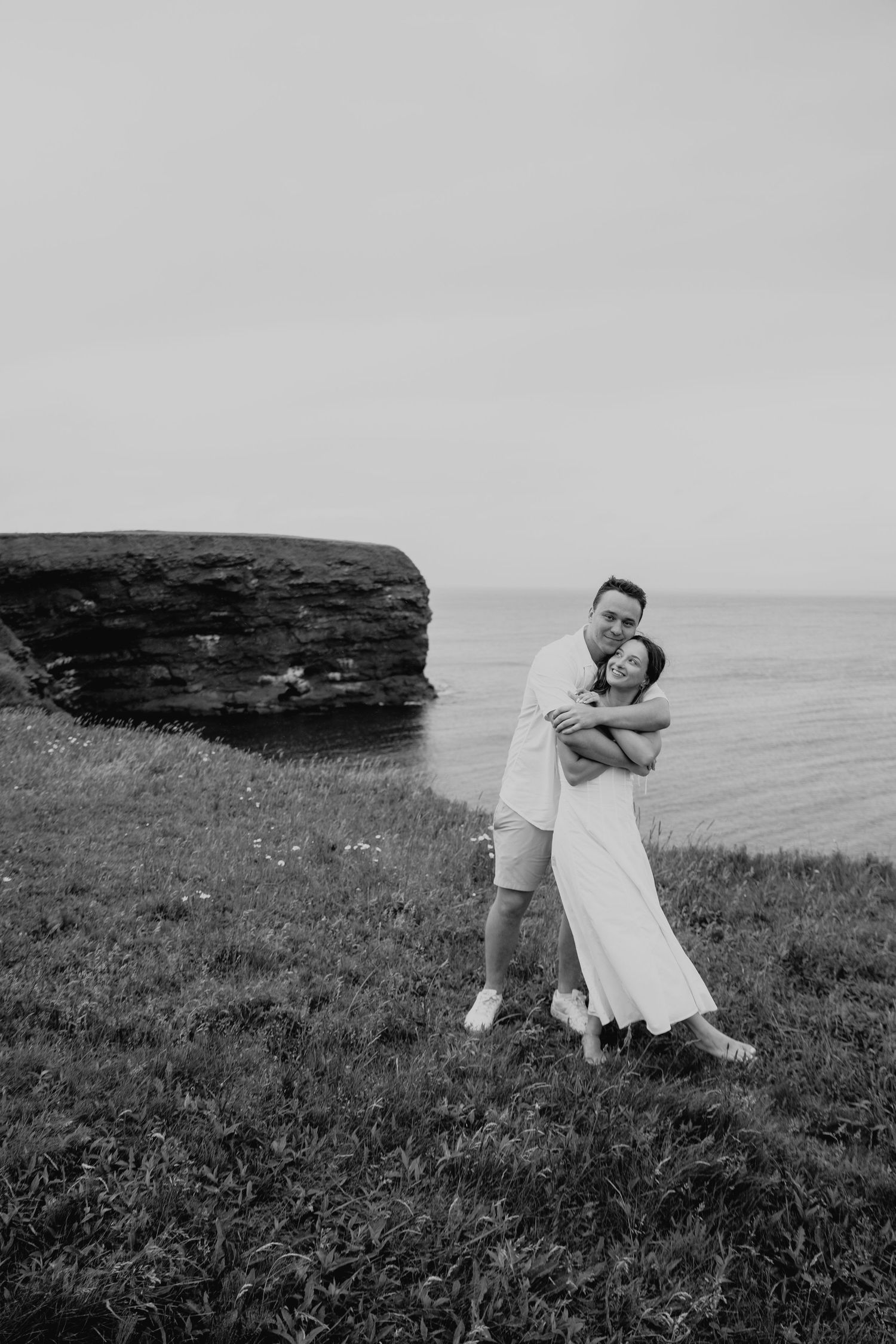 A romantic black and white photo series of a couple embracing on a grassy seaside cliff overlooking the ocean.