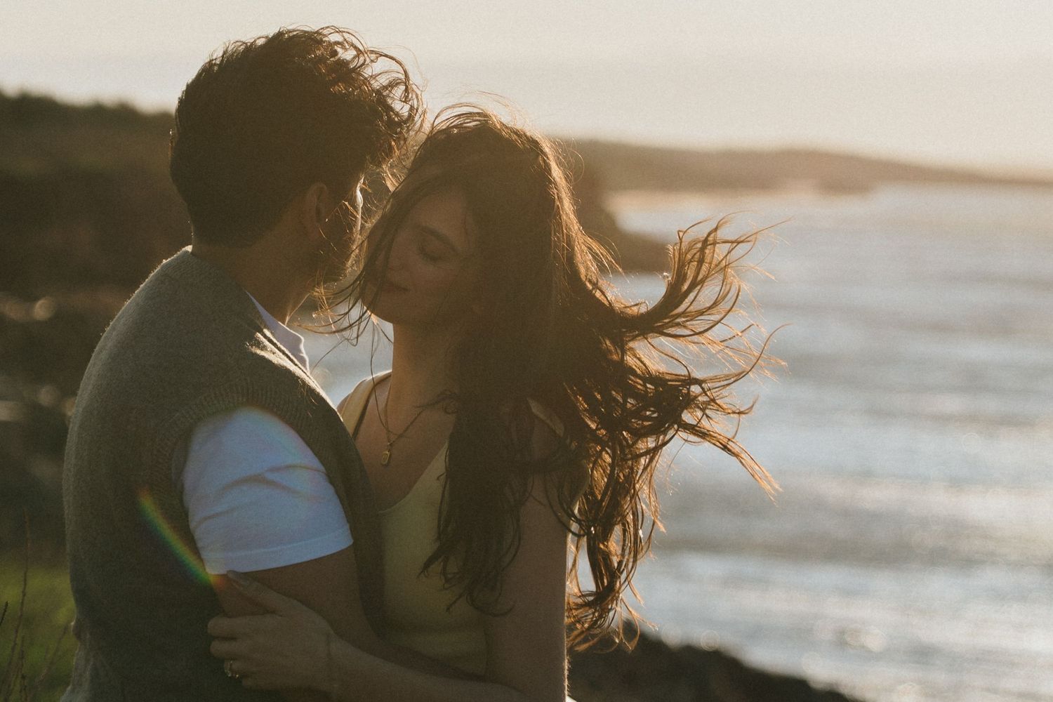 A couple embraces on a beach at sunset with wind blowing through their hair as ocean waves crash behind them.