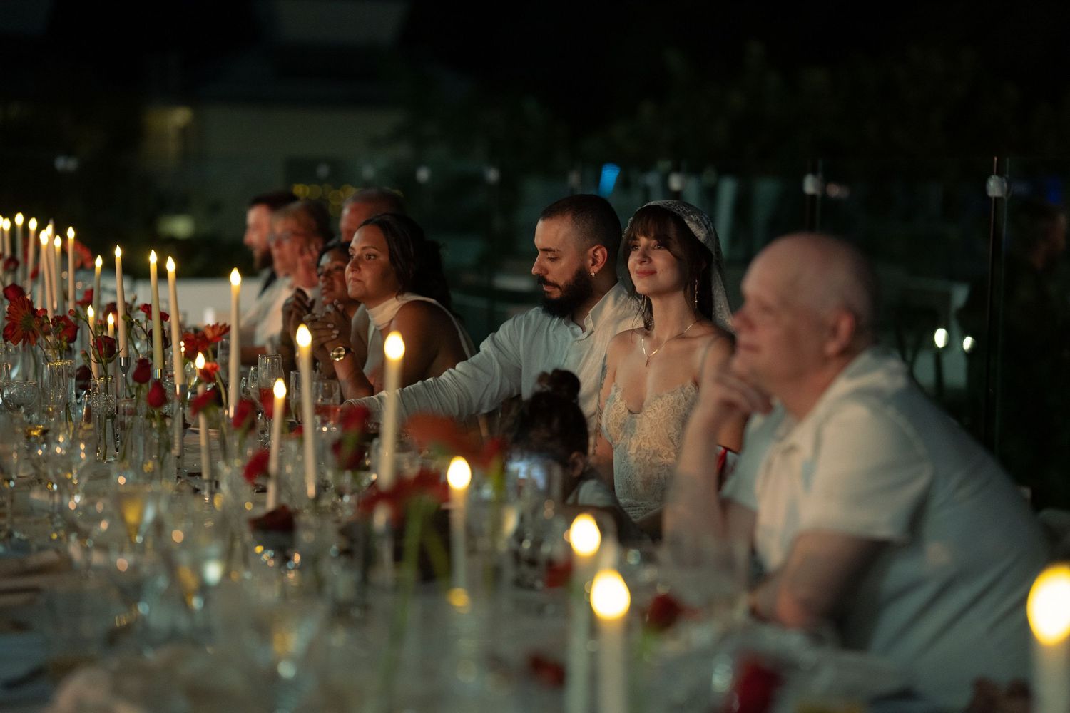 Wedding guests seated at candlelit reception dinner table at night with glowing candles and red rose decorations.