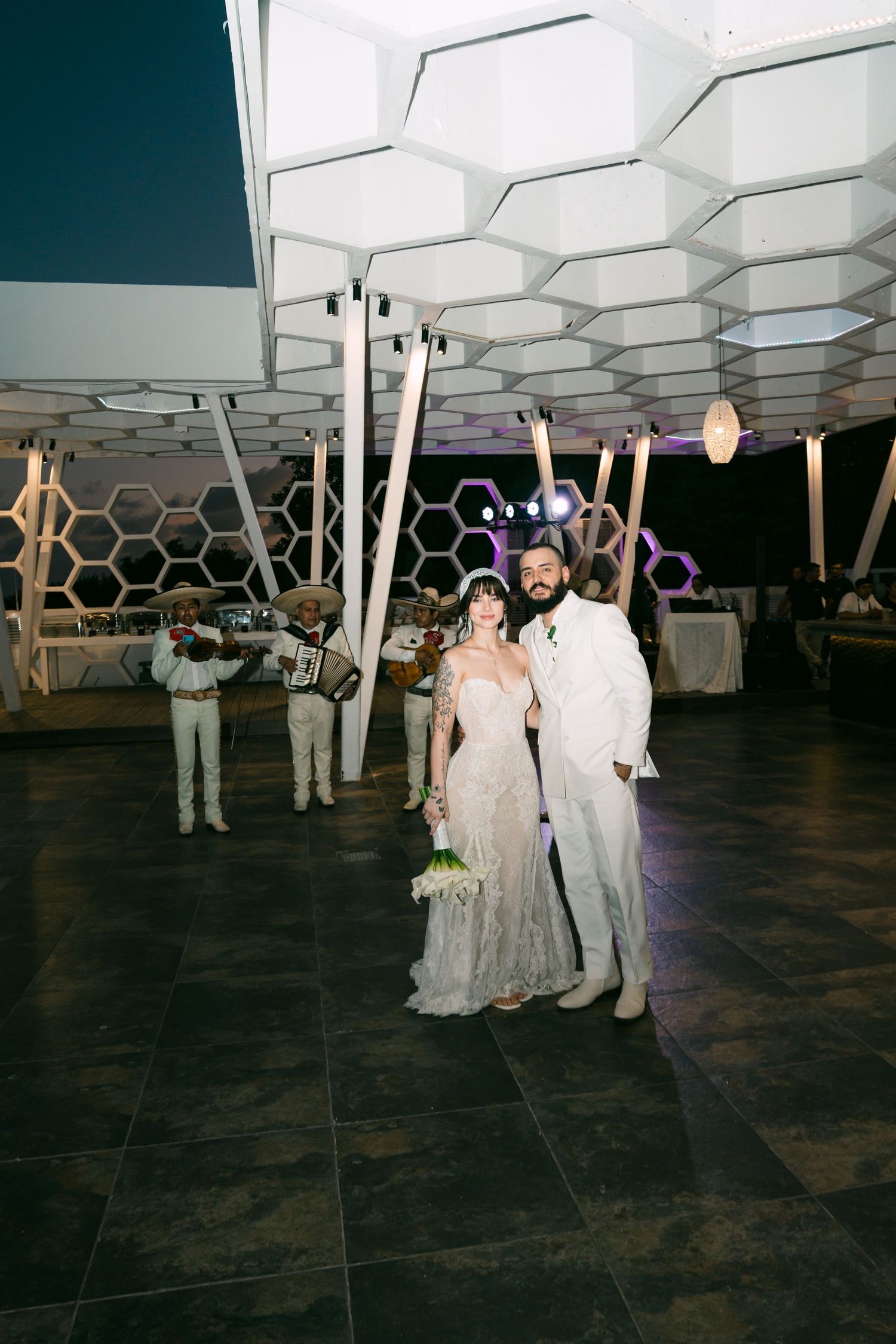 Bride and groom pose on dance floor at elegant outdoor wedding reception with honeycomb ceiling and mariachi band.