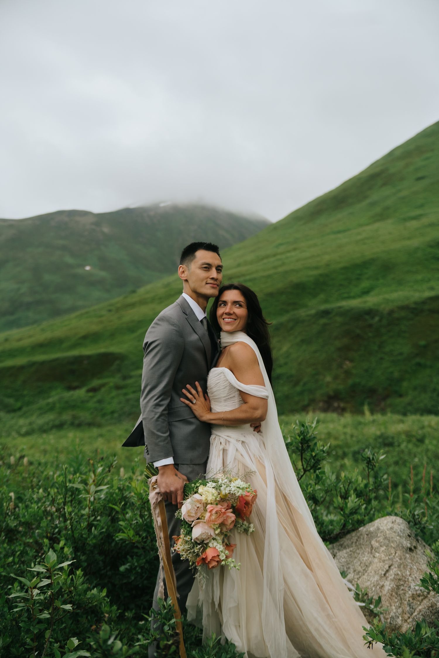 Newlyweds embrace on a misty mountainside surrounded by verdant landscape in Alaska