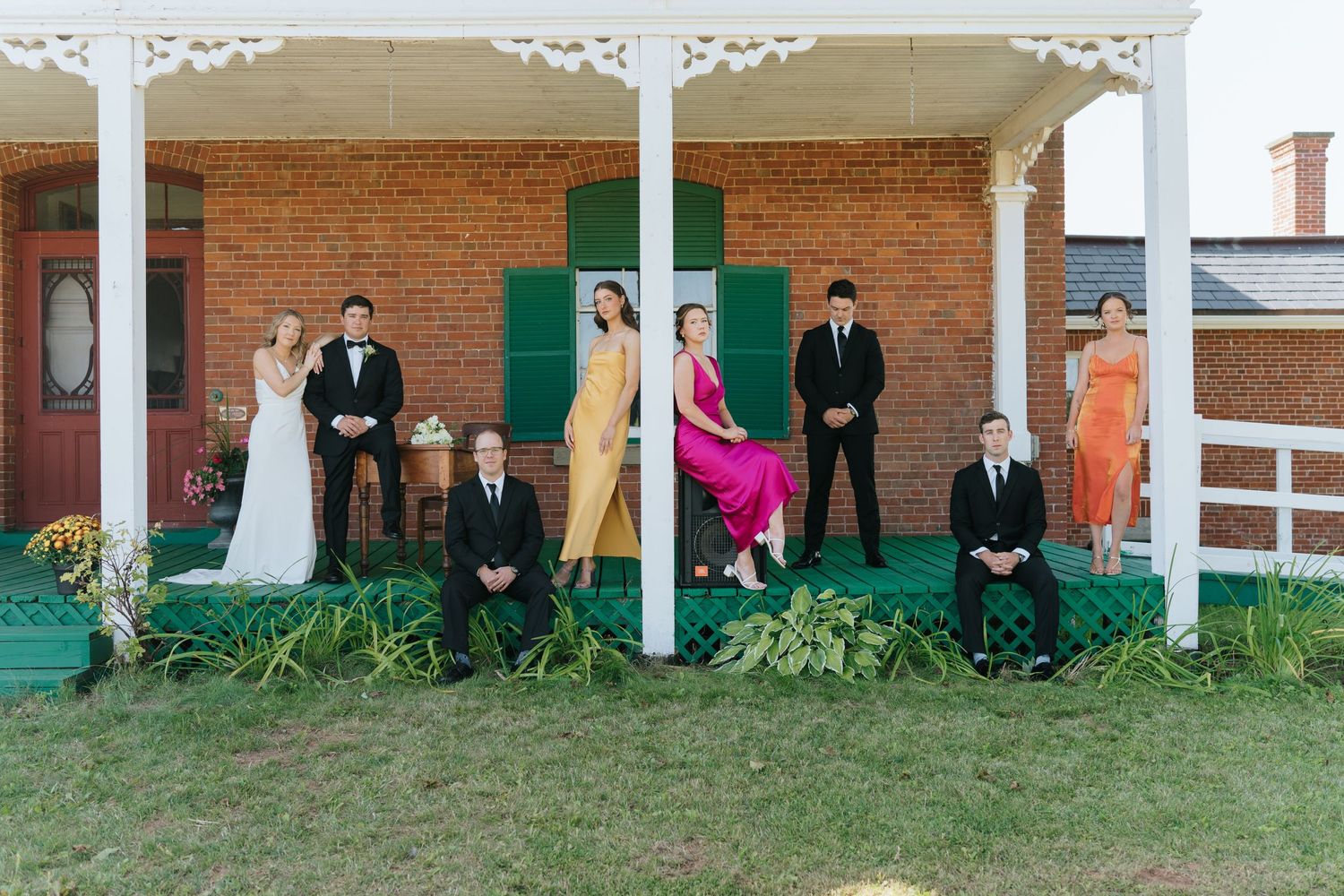Wedding party members stand in a line against a brick building with white columns and green trim.