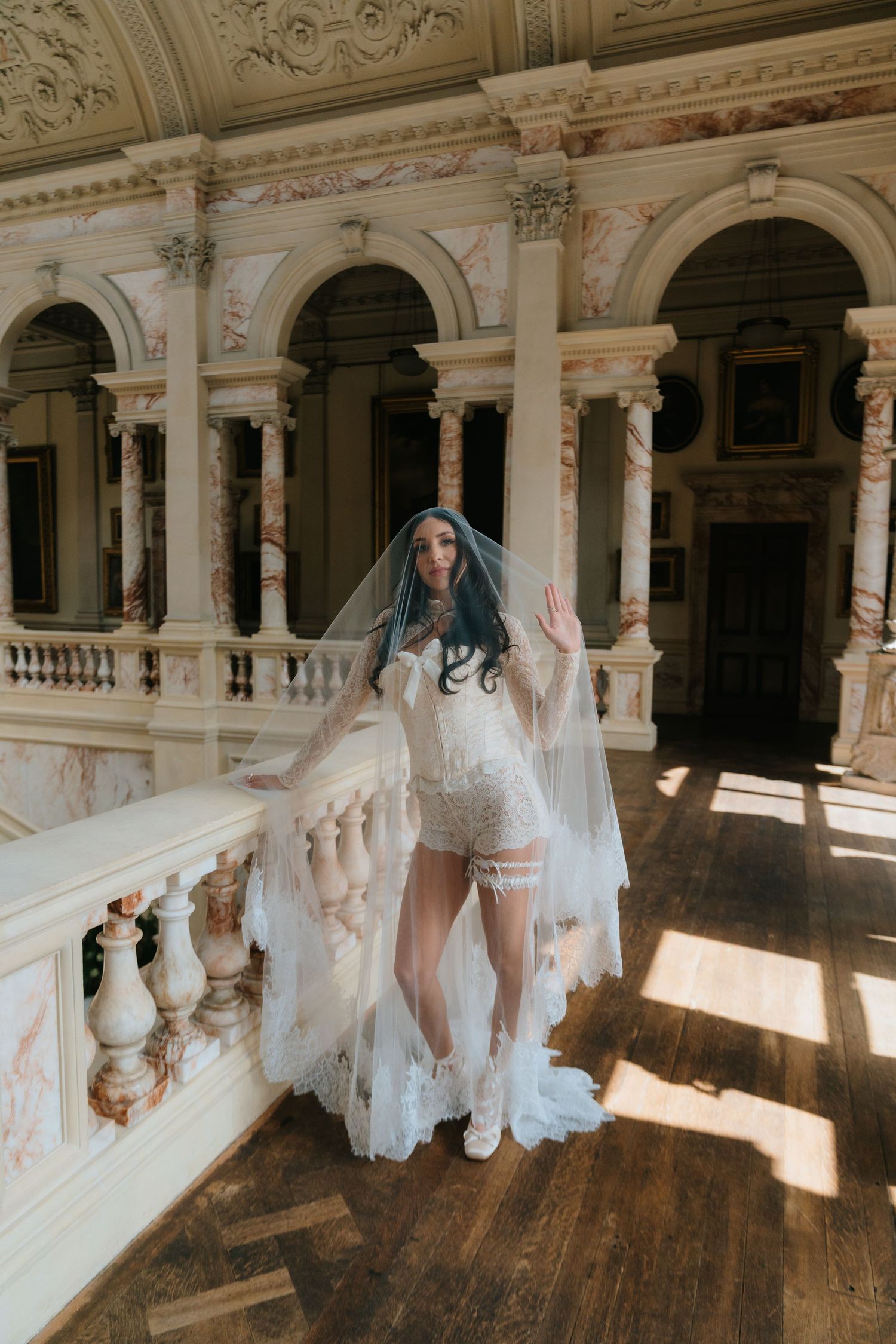 Bride in white lace corset and flowing veil stands on a grand marble balcony in a luxurious historic mansion.
