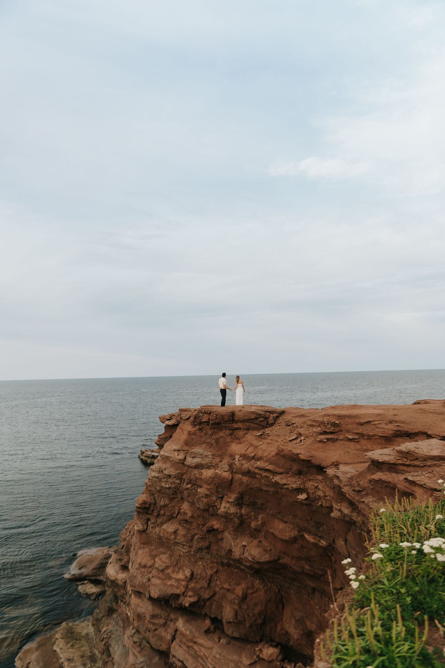 Two figures stand on dramatic red cliffs overlooking the ocean at sunset along a rugged coastline.