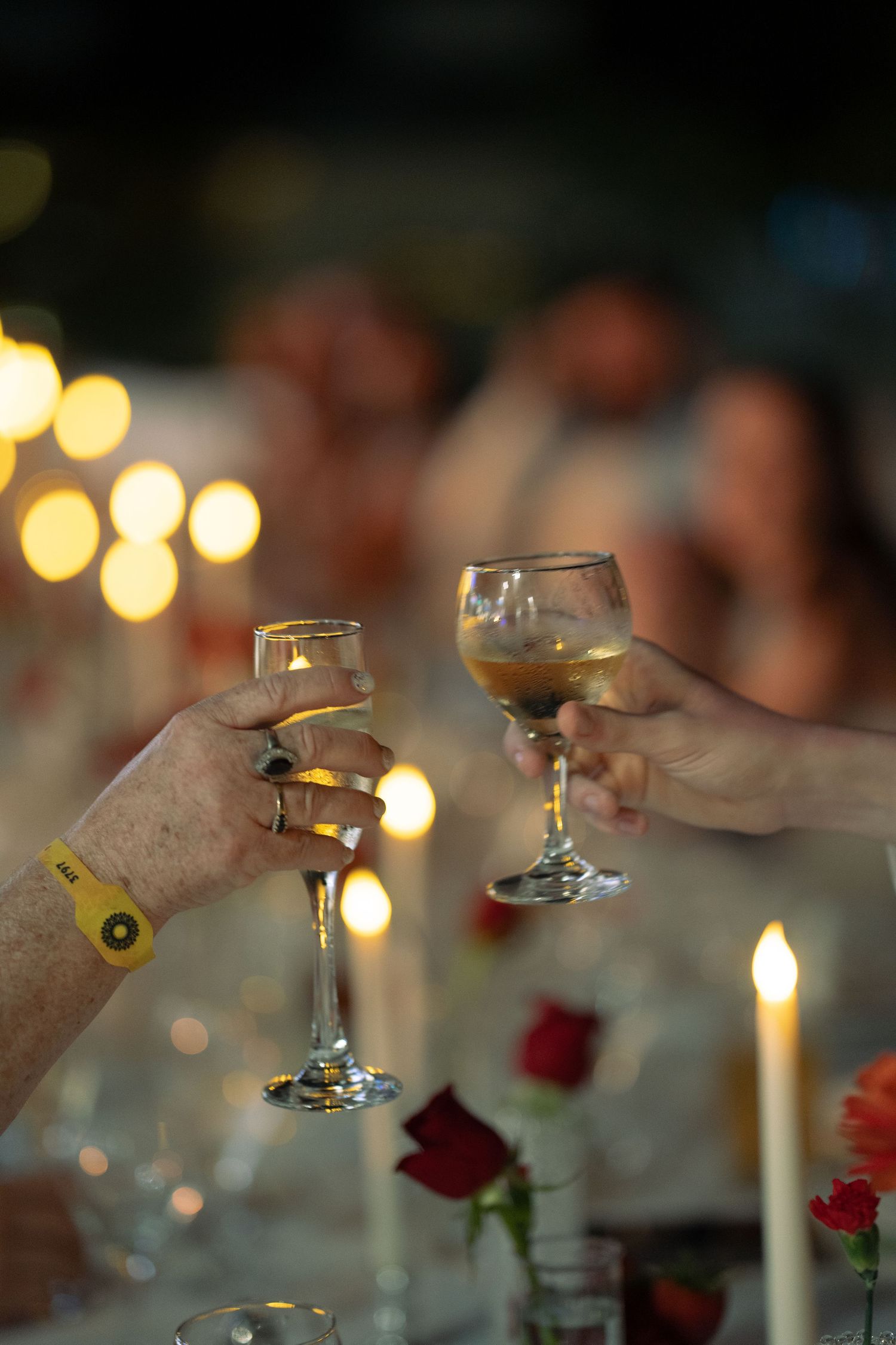 Two hands clinking wine glasses at a candlelit dinner party with roses and bokeh lights in background.
