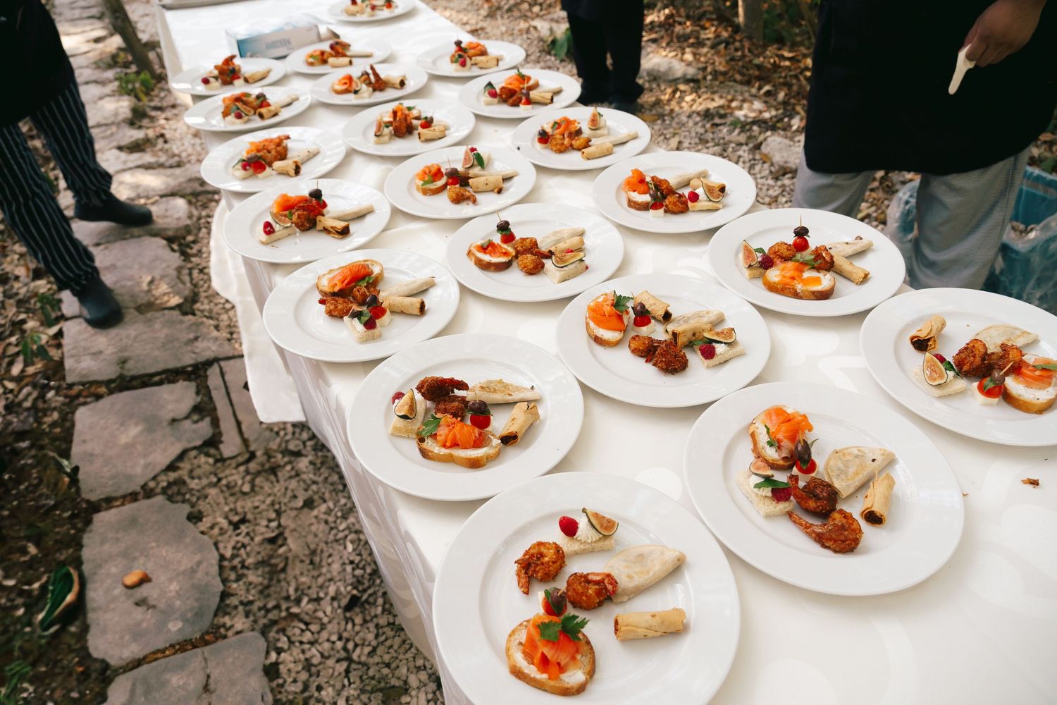 Rows of elegant white plates with gourmet appetizers displayed on a white tablecloth at an outdoor catering event.